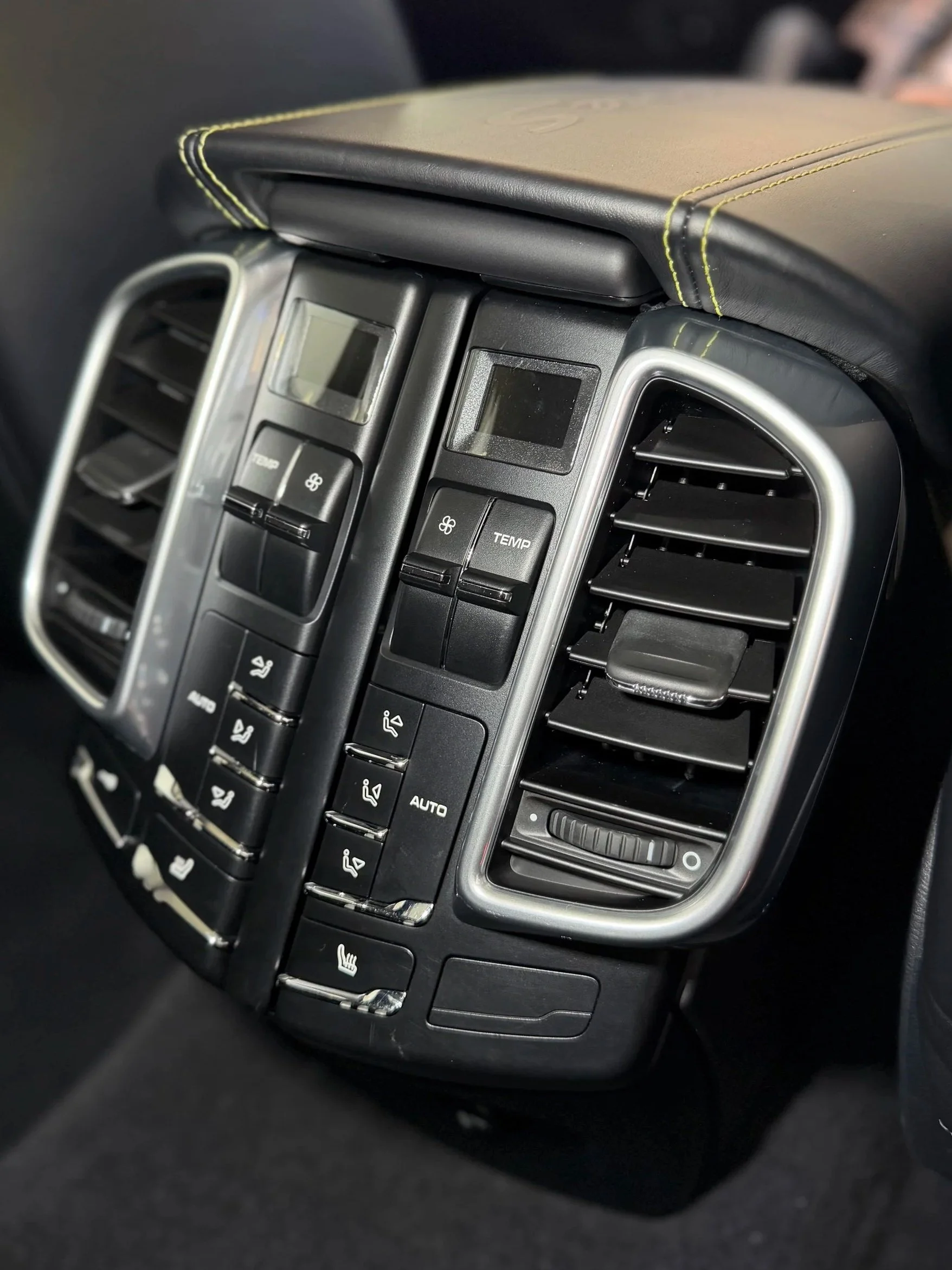 Close-up of a car's center console with air vents, climate control buttons, and a brown leather pad with yellow stitching on top. Cleaned air vents