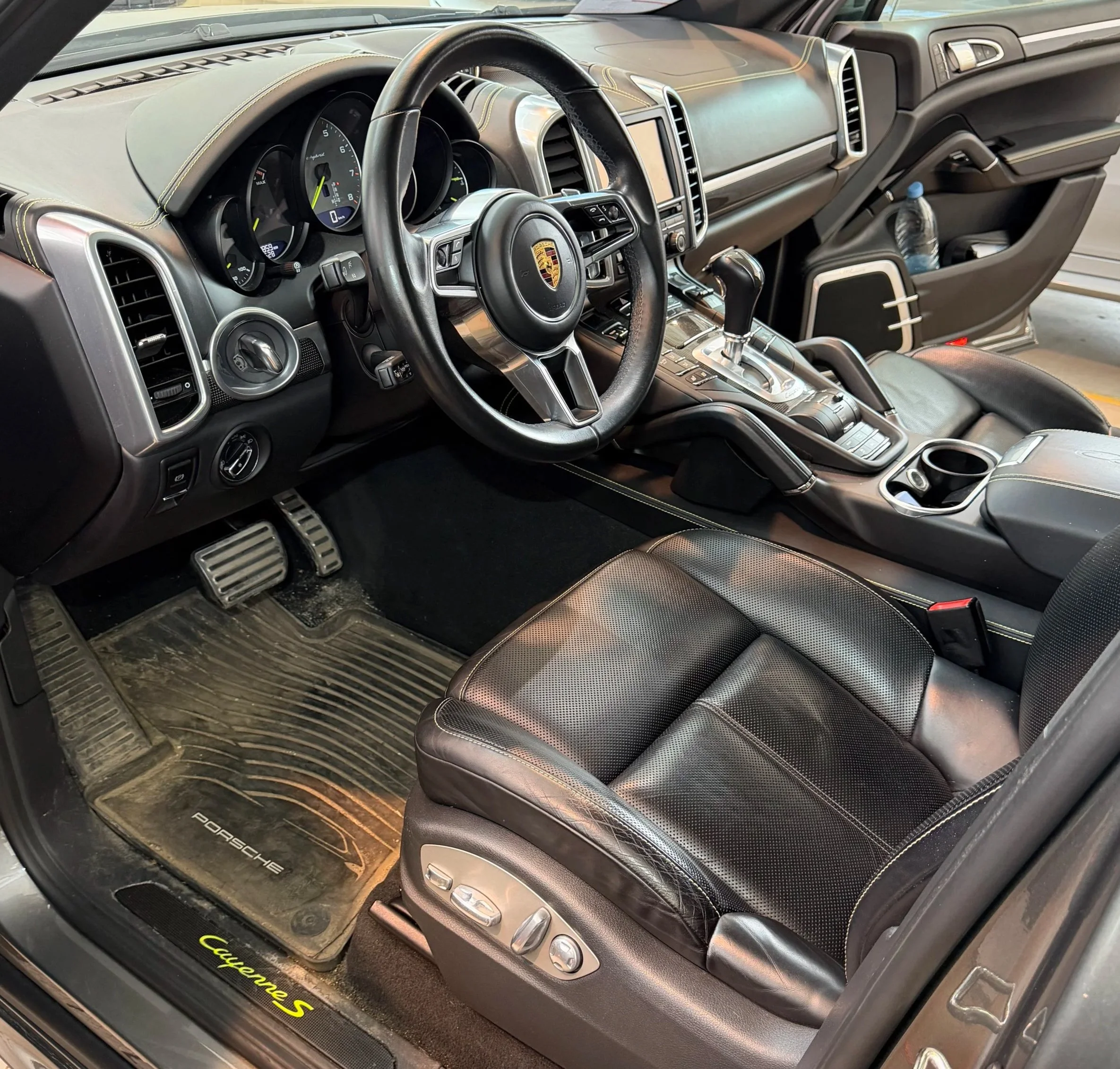 The interior of a luxury Porsche Cayenne S sports vehicle, focusing on the steering wheel, dashboard, center console, and black leather seats. Dirty interior