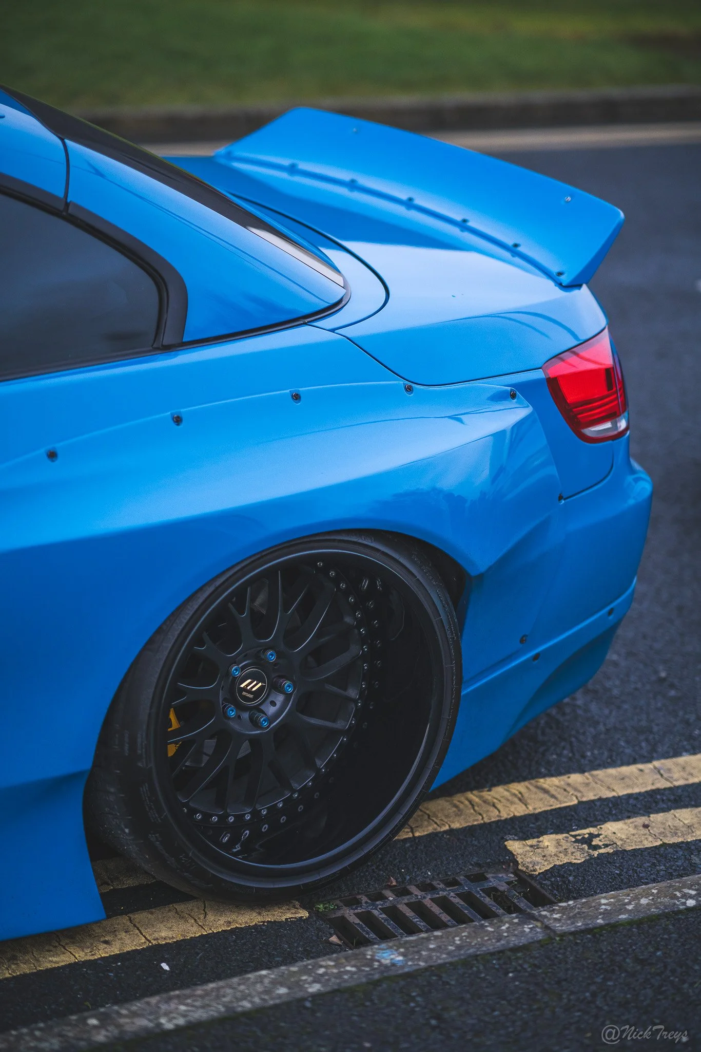 Close-up of a blue sports car's rear section, showcasing a large rear wing spoiler, black aftermarket wheel, and part of the rear taillight, parked on a street. BMW 335i pandem kit