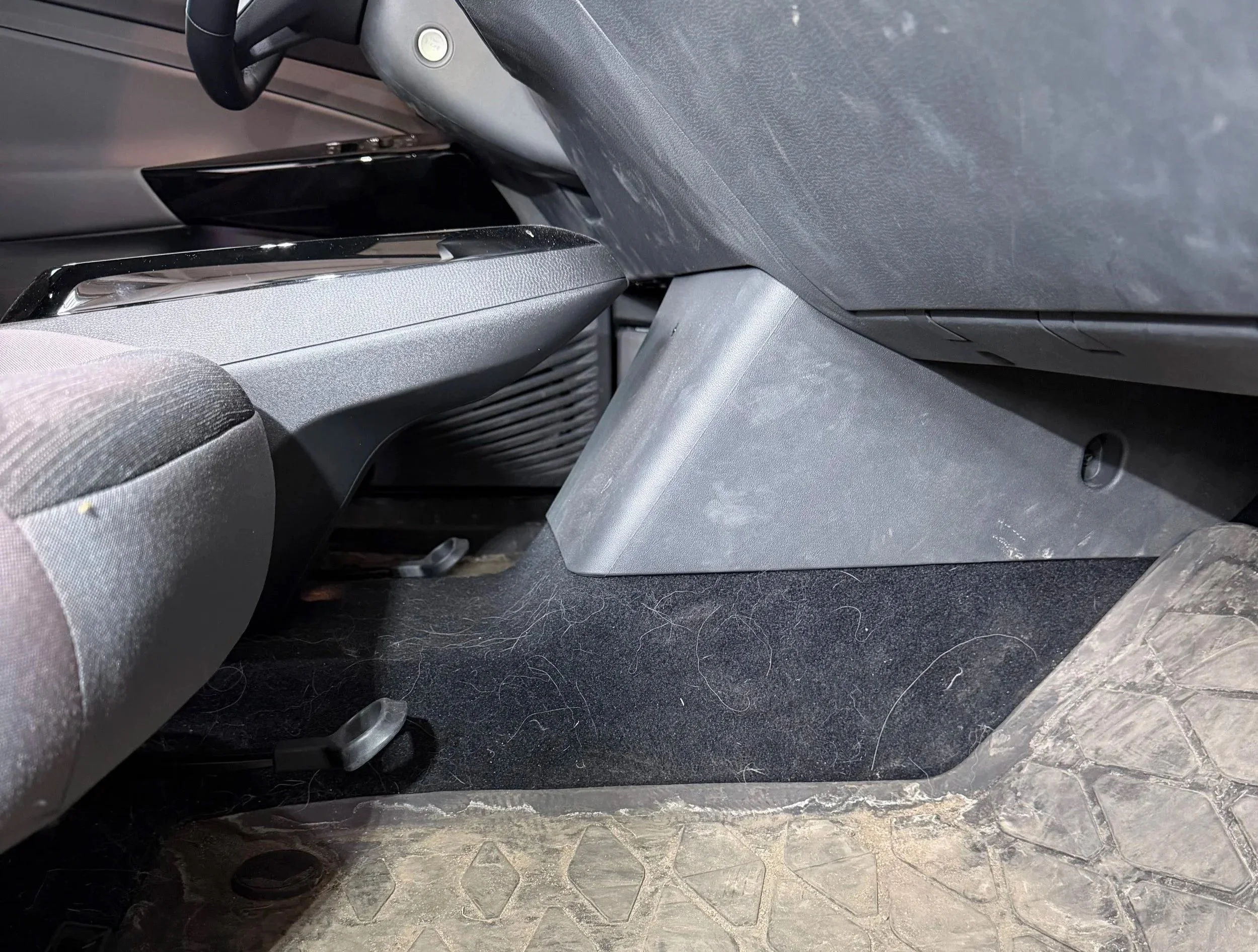 The interior of a vehicle showing the area below the dashboard, with visible pedals on the floor, including accelerator and brake pedals, and part of the driver's side seat and carpeted floor mat. Dirty interior with pet hair
