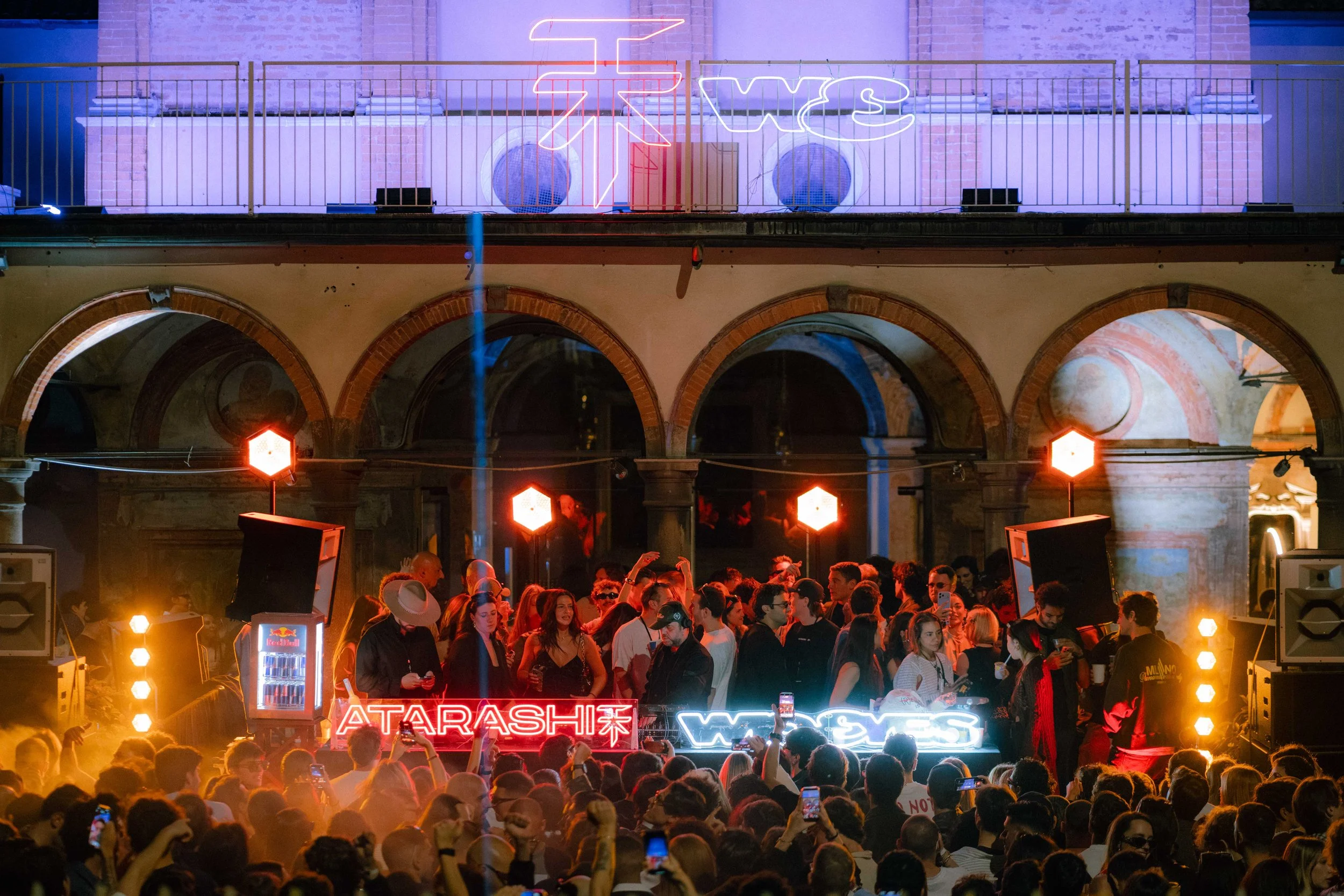 Una scena di festa con molte persone radunate in un ambiente urbano con archi di mattoni. Sullo sfondo, luci al neon con parole come 'WES', 'ATARASHI', e 'RED BULL'. La gente sta ballando e si diverte sotto luci calde e rosse.