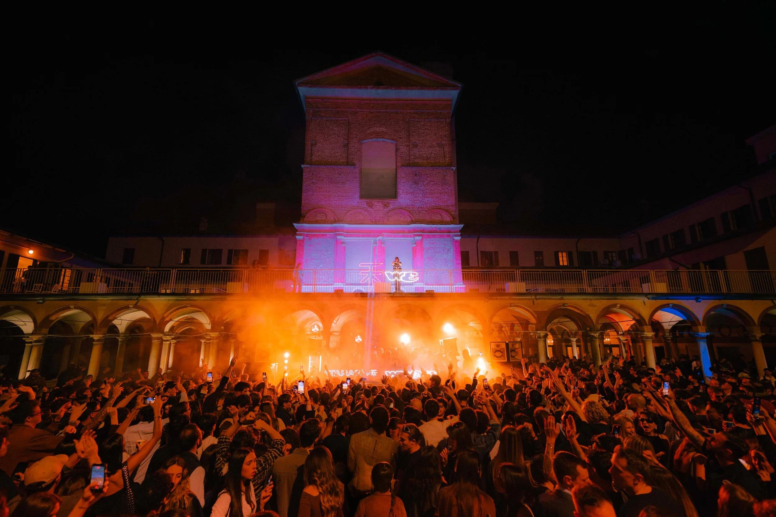 Un grande evento con una folla di persone che guarda un palco illuminato con luci colorate e una struttura storica sullo sfondo, decorata con luci e proiezioni, durante la notte.