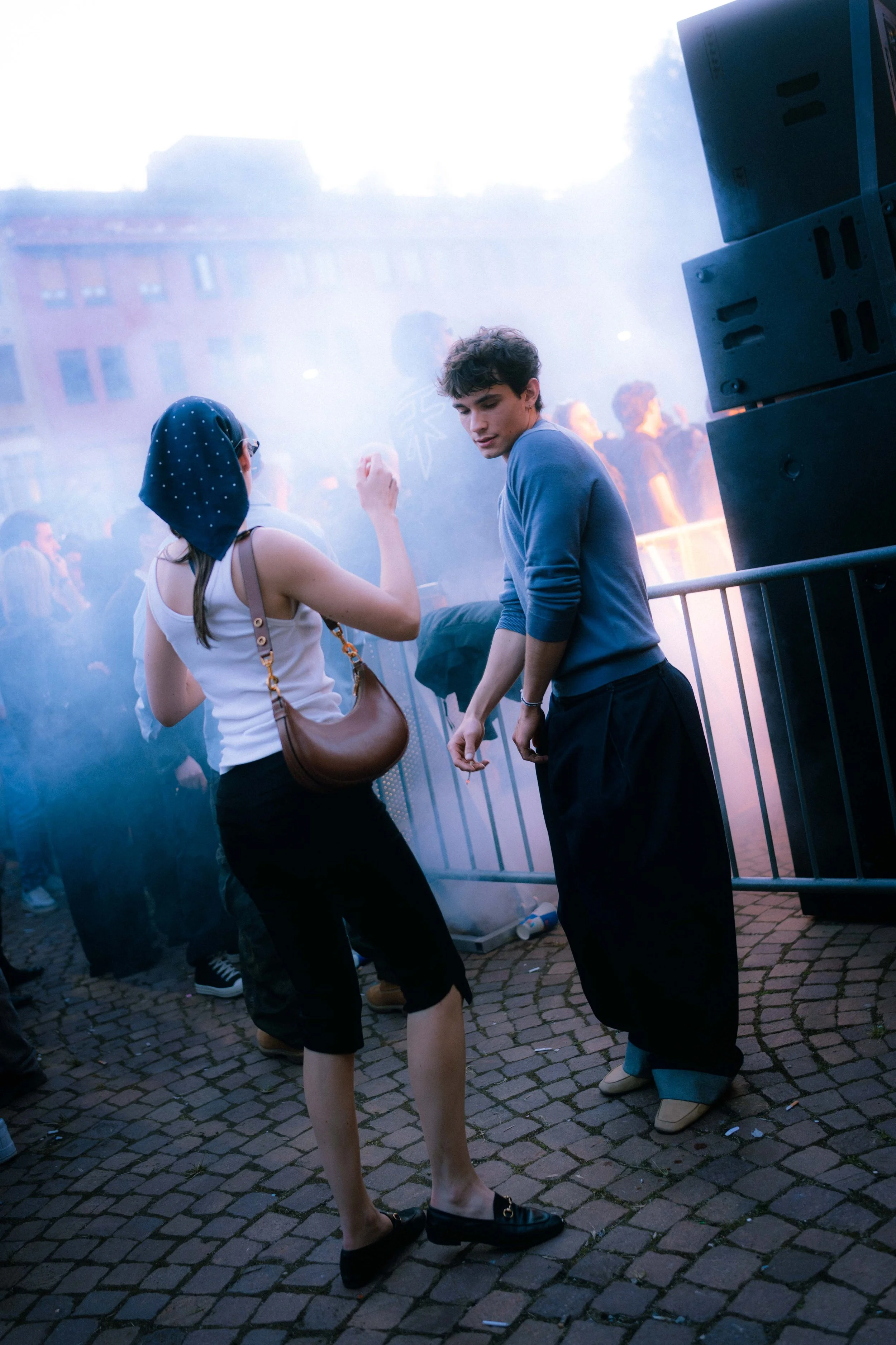 Due persone, una donna con vestiti bianchi e una borsa marrone e un uomo con pantaloni neri e maglietta blu, stanno danzando tra la folla sotto un cielo nuvoloso, circondati da fumo e musica in un evento all'aperto.