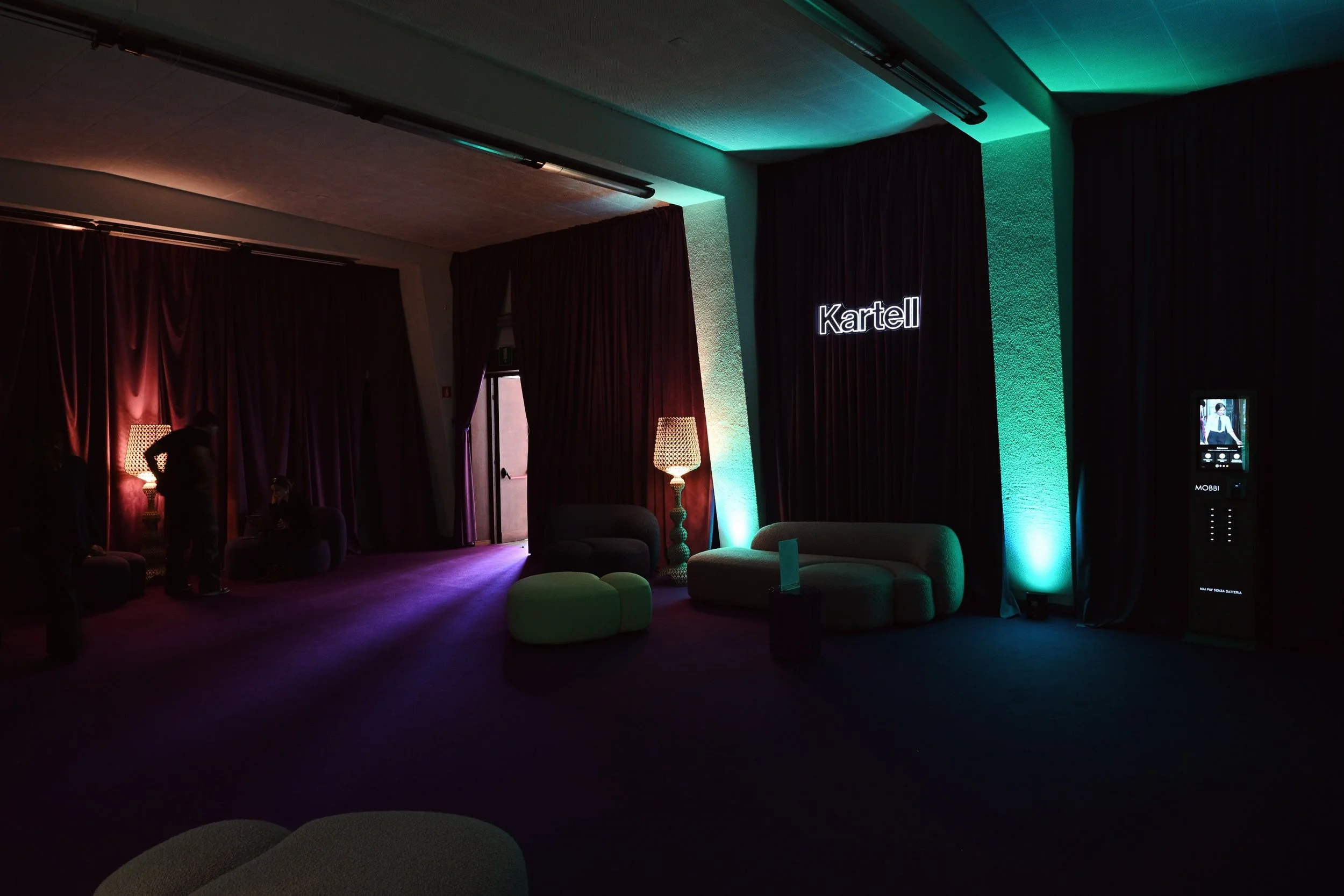 Interno di un locale con luci soffuse, divani verdi e tende viola. Una lampada luminosa e un cartello con il nome "Kartell" sono visibili.