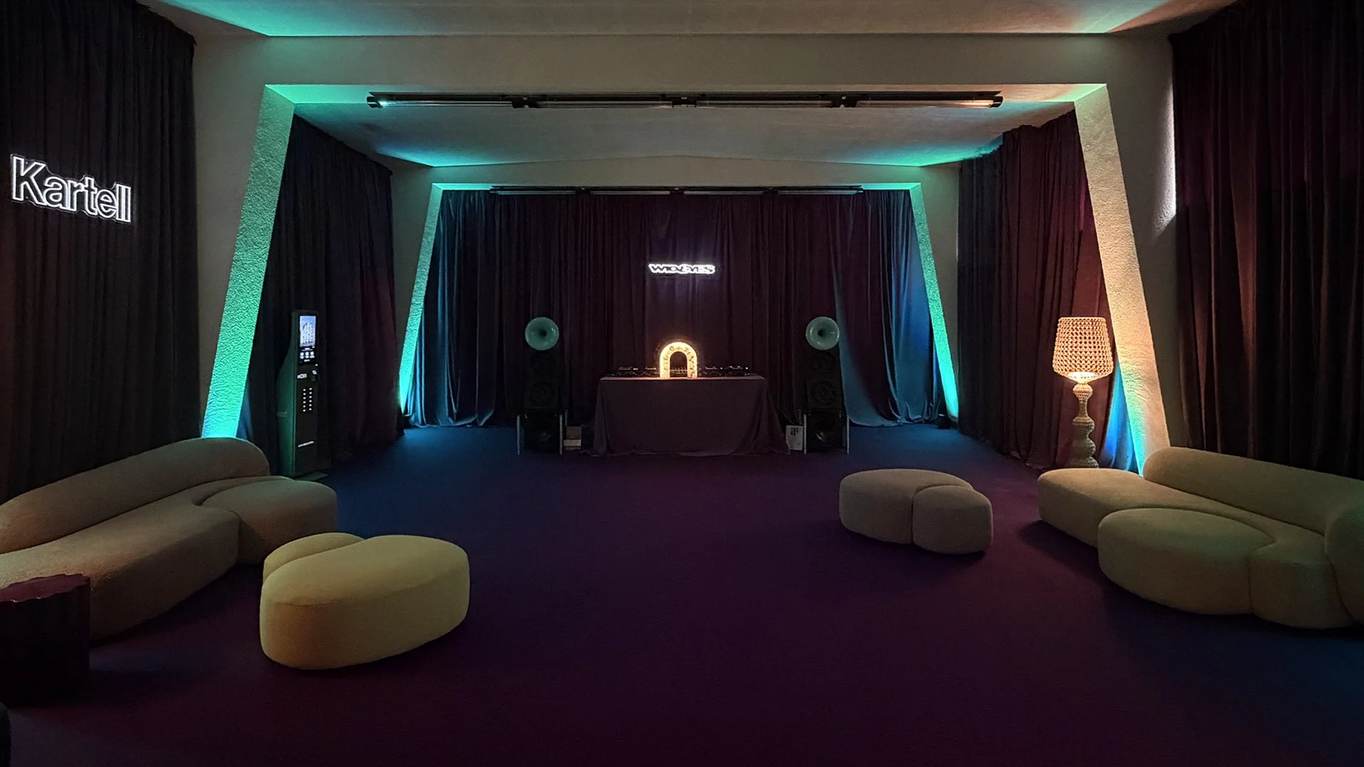 Sala con luci colorate, divani, pouf, lampada e arredamento in stile rétro, con palco e decorazioni per eventi o musica dal vivo.
