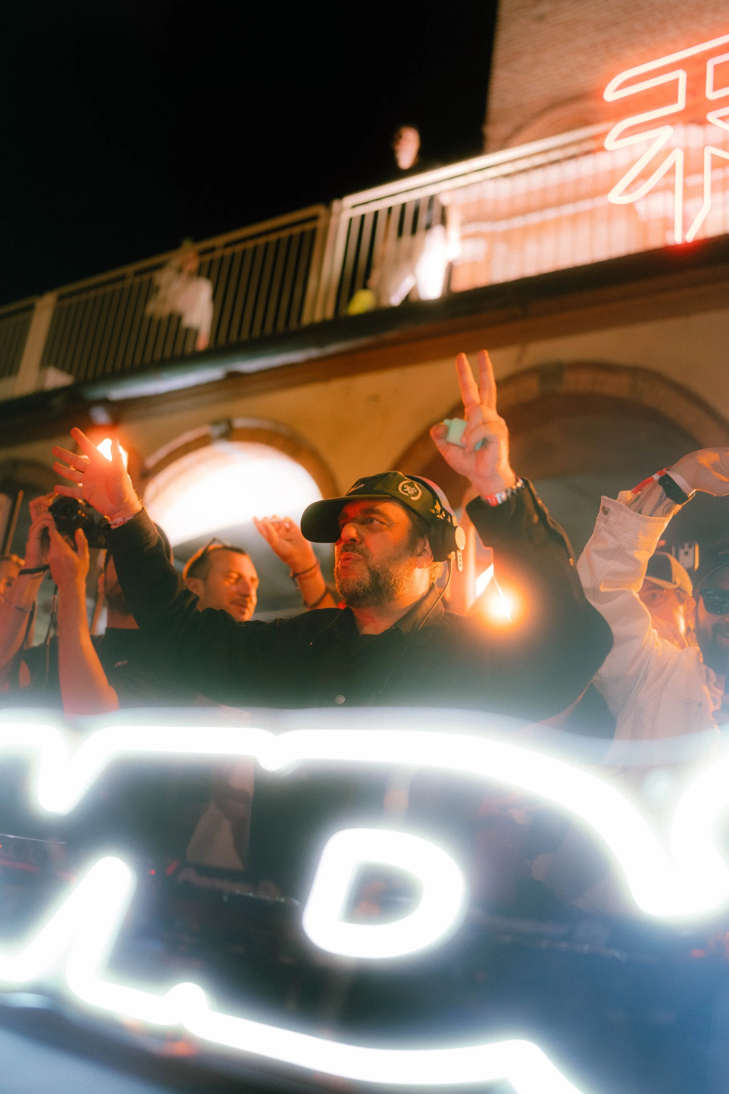 Persone che festeggiano e fanno feste notturne, alcune mostrano il segno della pace con le dita, in un ambiente illuminato da luci neon e con un'insegna luminosa.