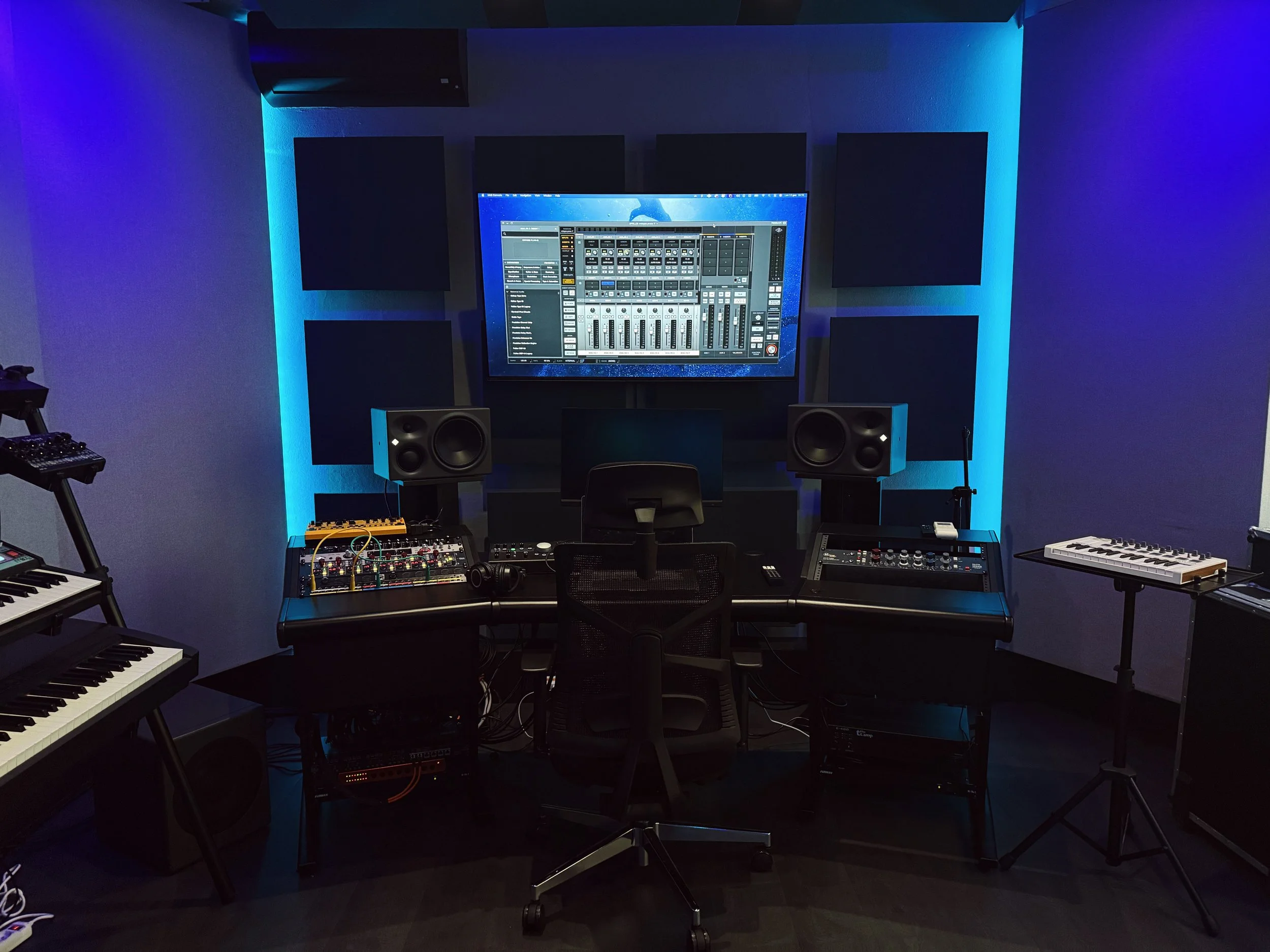 Stanza di produzione musicale con schermo, altoparlanti, tastiere e strumenti elettronici, illuminata da luci blu.
