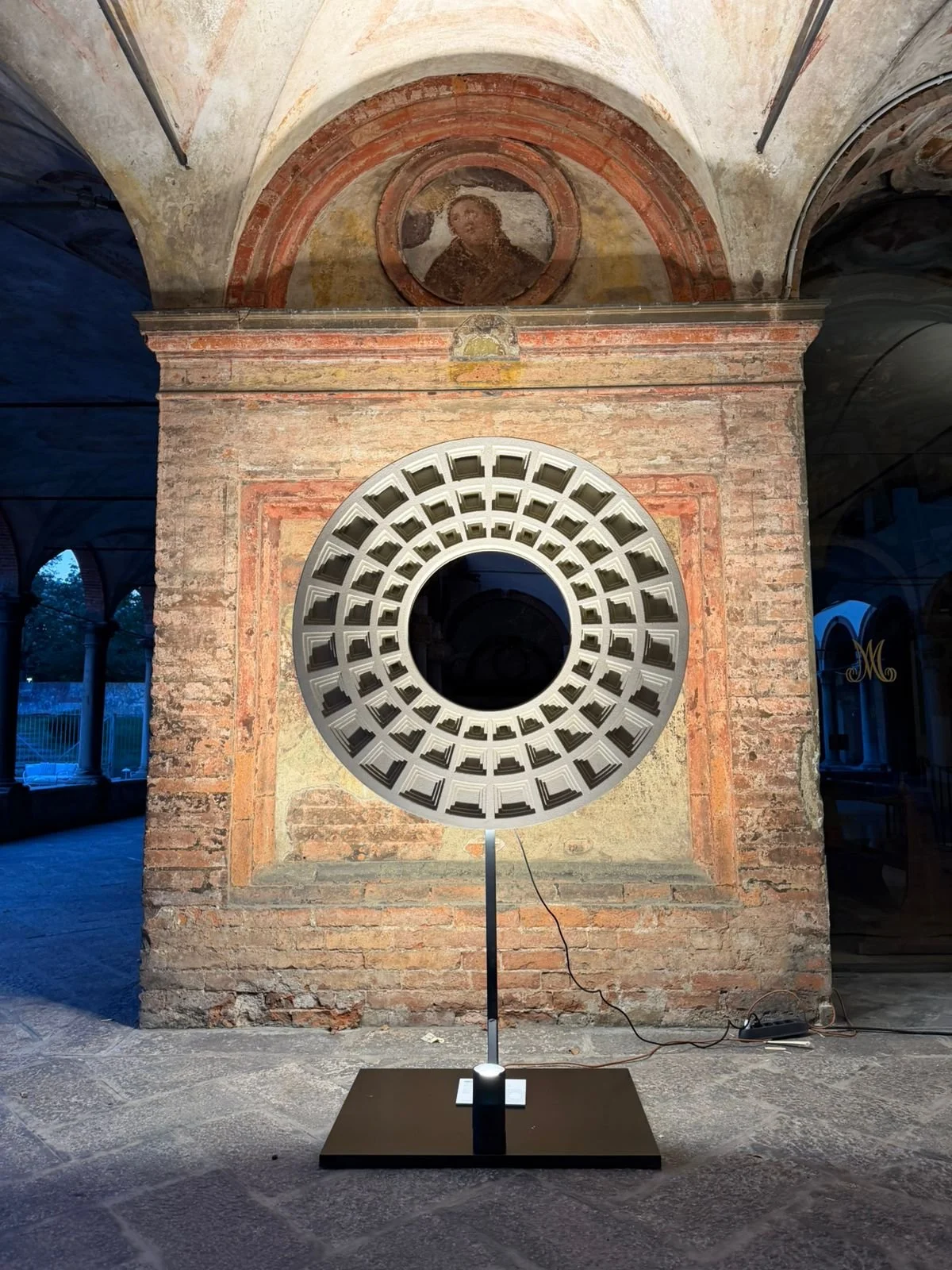 Installazione artistica con grande cerchio di luci in un ambiente storico, con pareti di mattoni e affreschi.