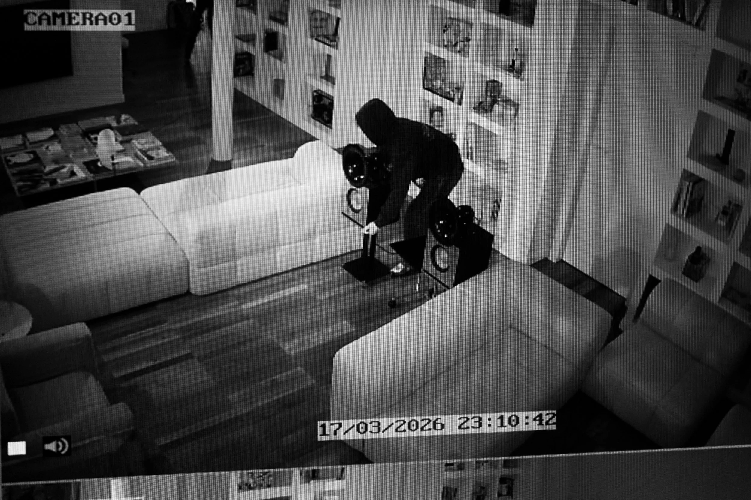 Una scena di sorveglianza notturna che mostra una persona con cappuccio e cappello nero che si piega su un divano bianco, vicino a due grandi altoparlanti neri. Sullo sfondo, scaffali con libri e oggetti decorativi. Data e ora visibili sullo schermo: 17/03/2026 23:10:42.