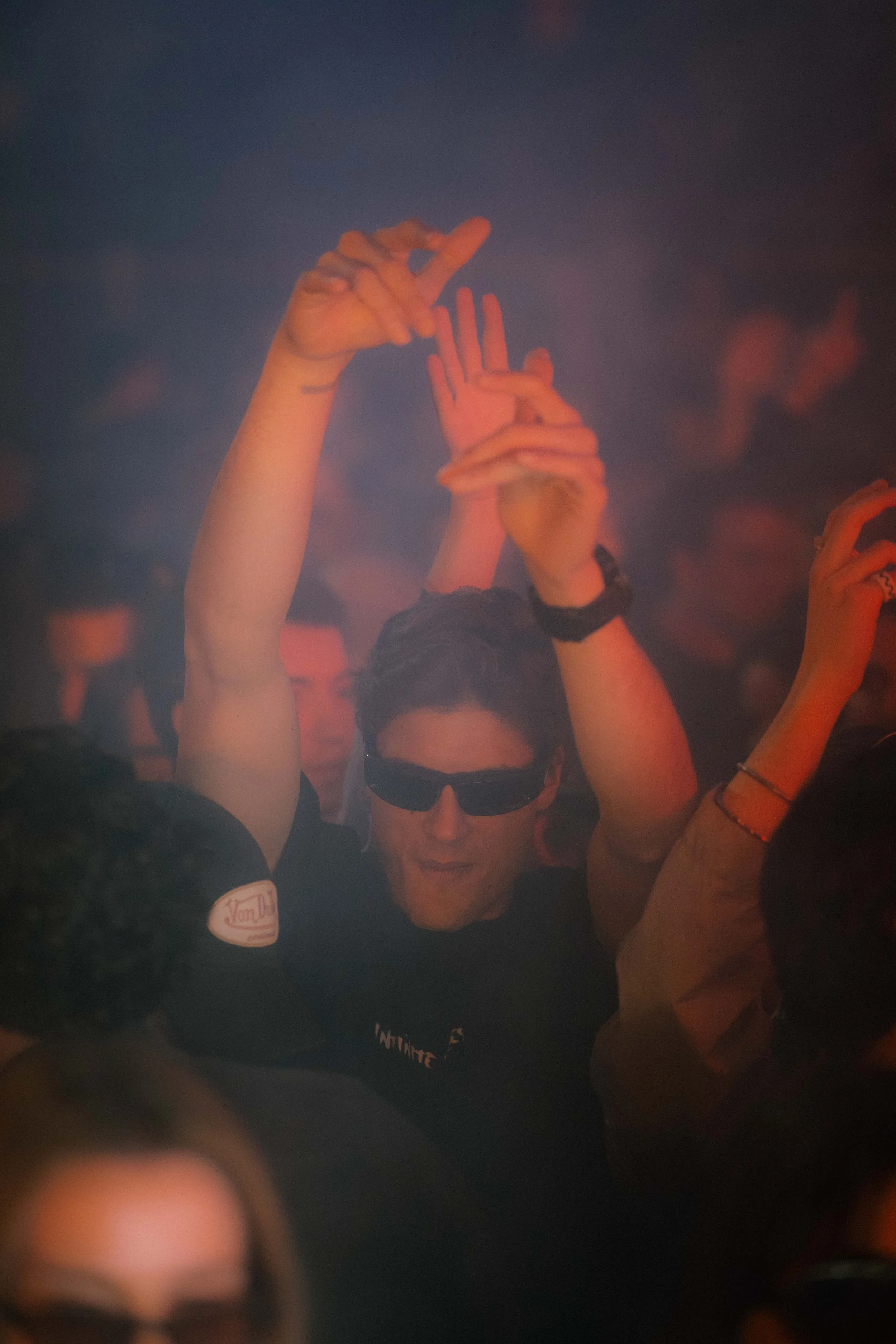 Immagine di una persona con occhiali da sole che alza le mani in un ambiente oscuro e affollato, presumibilmente in una discoteca o a un concerto, con altre persone sullo sfondo.