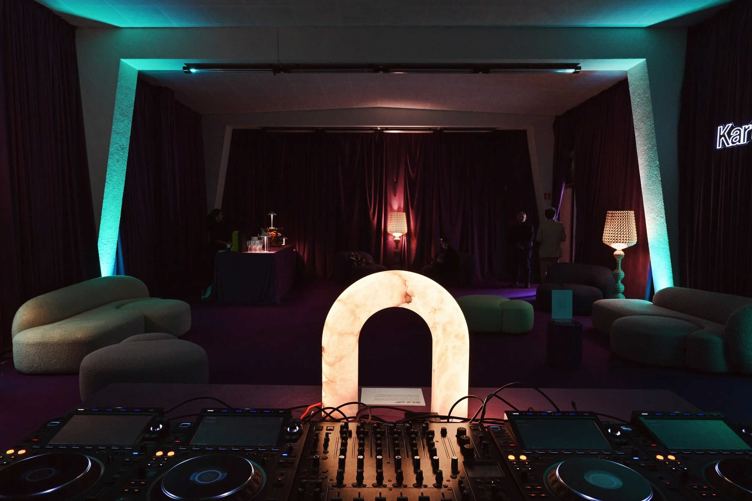 Interno di un nightclub con luci soffuse, grandi lampade da tavolo, divani color crema, una pista da ballo con un palco con una tenda rossa e persone che parlano. Al centro un DJ set.