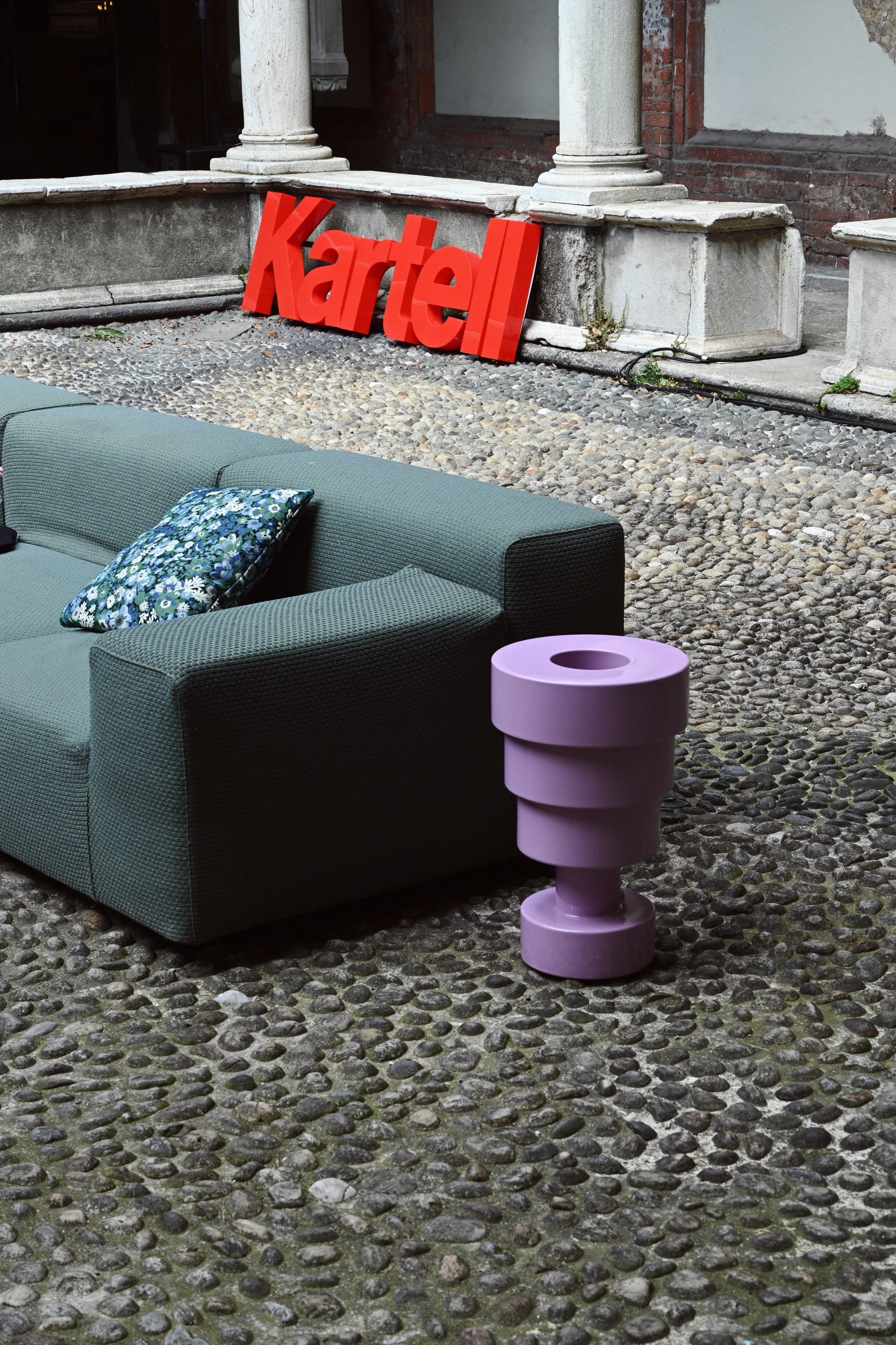Arredamento esterno con divano verde, cuscino floreale, tavolo viola, decorazioni in pietra e pareti di mattoni e pietra