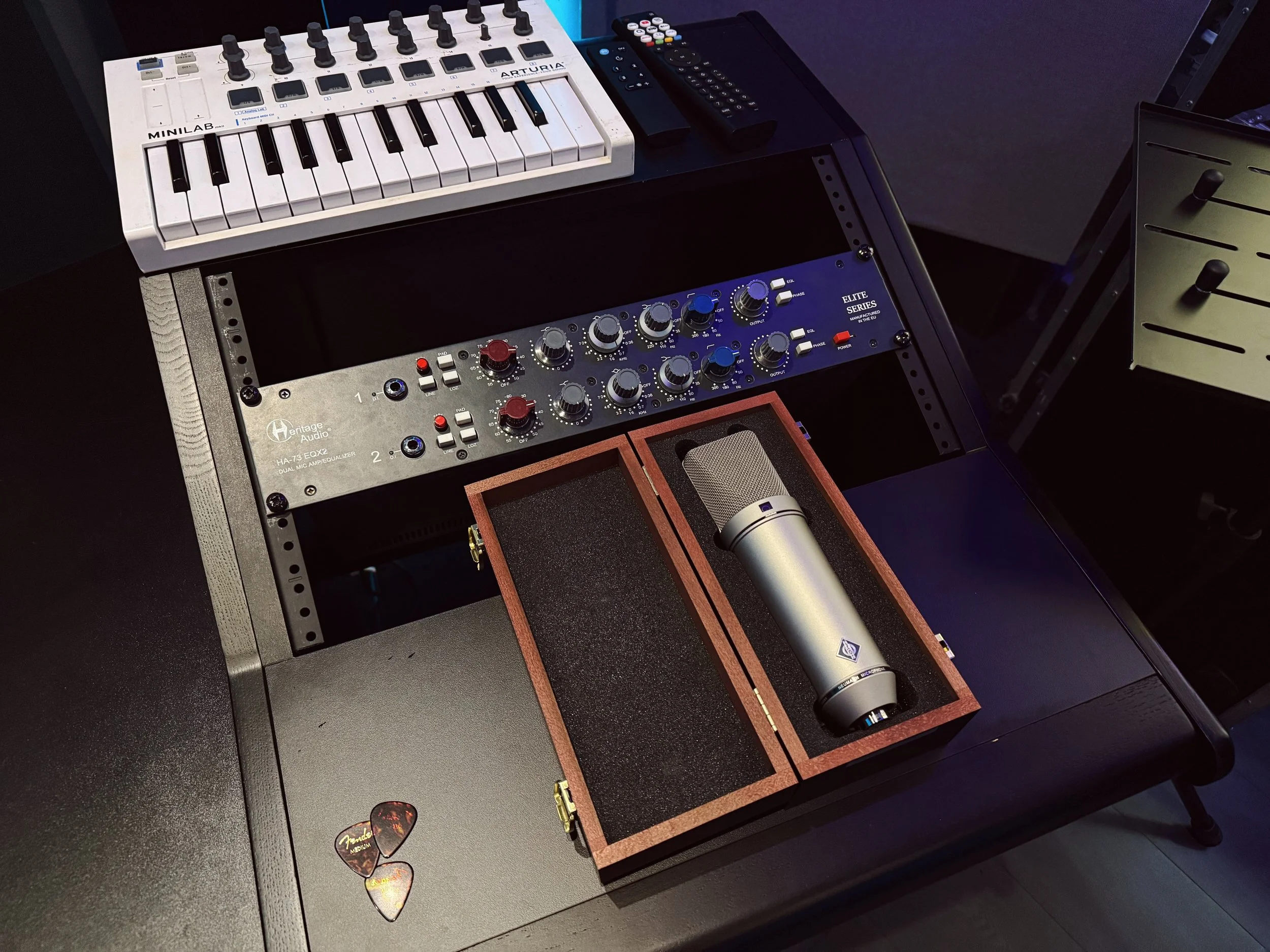 Immagine di un setup musicale con un sintetizzatore, un microfono in una scatola di legno, un equalizzatore e vari telecomandi e oggetti su una superficie.