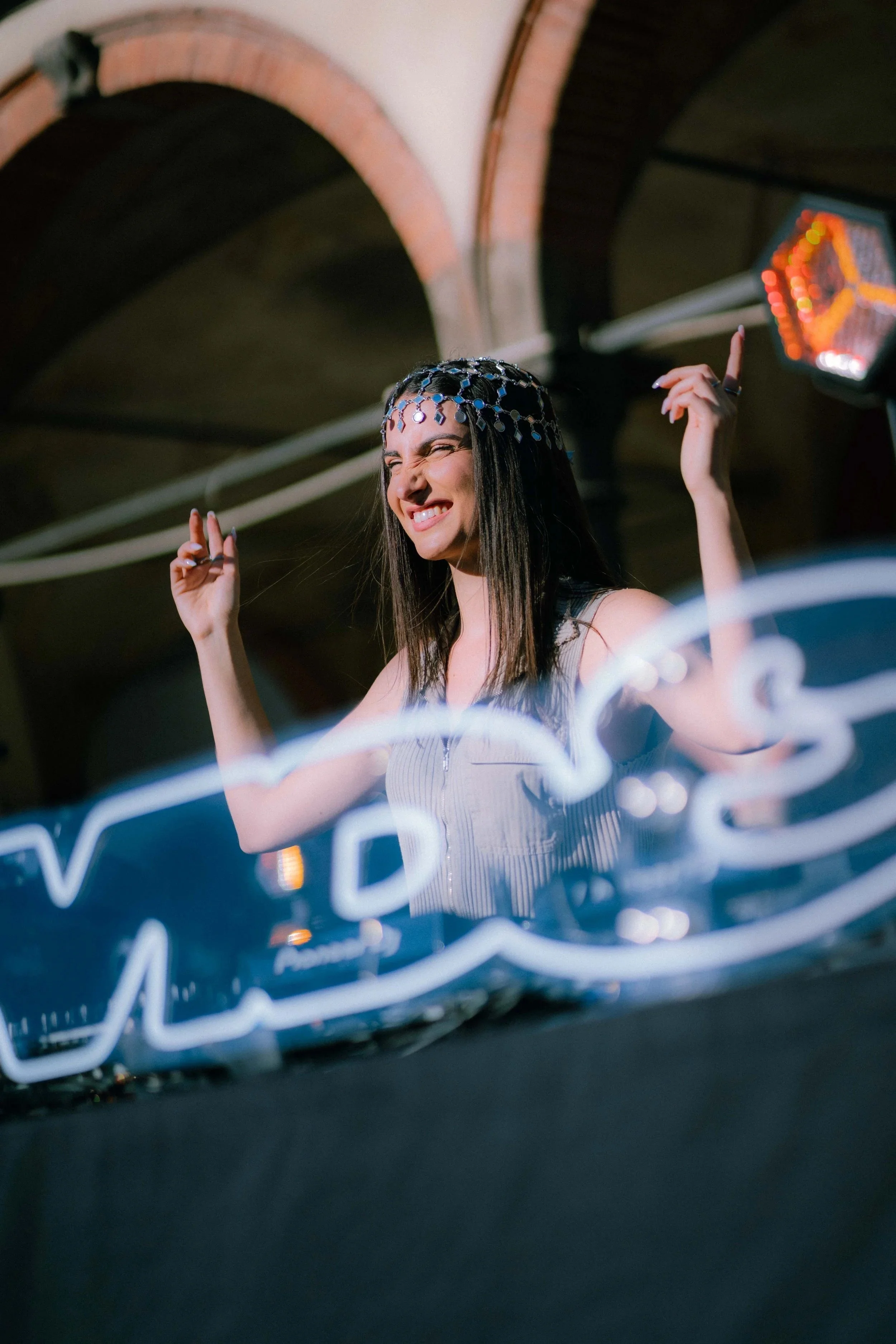 Una donna sorridente con una corona di luci sta pendant alla consolle di un DJ, con un ambiente notturno e luci colorate.