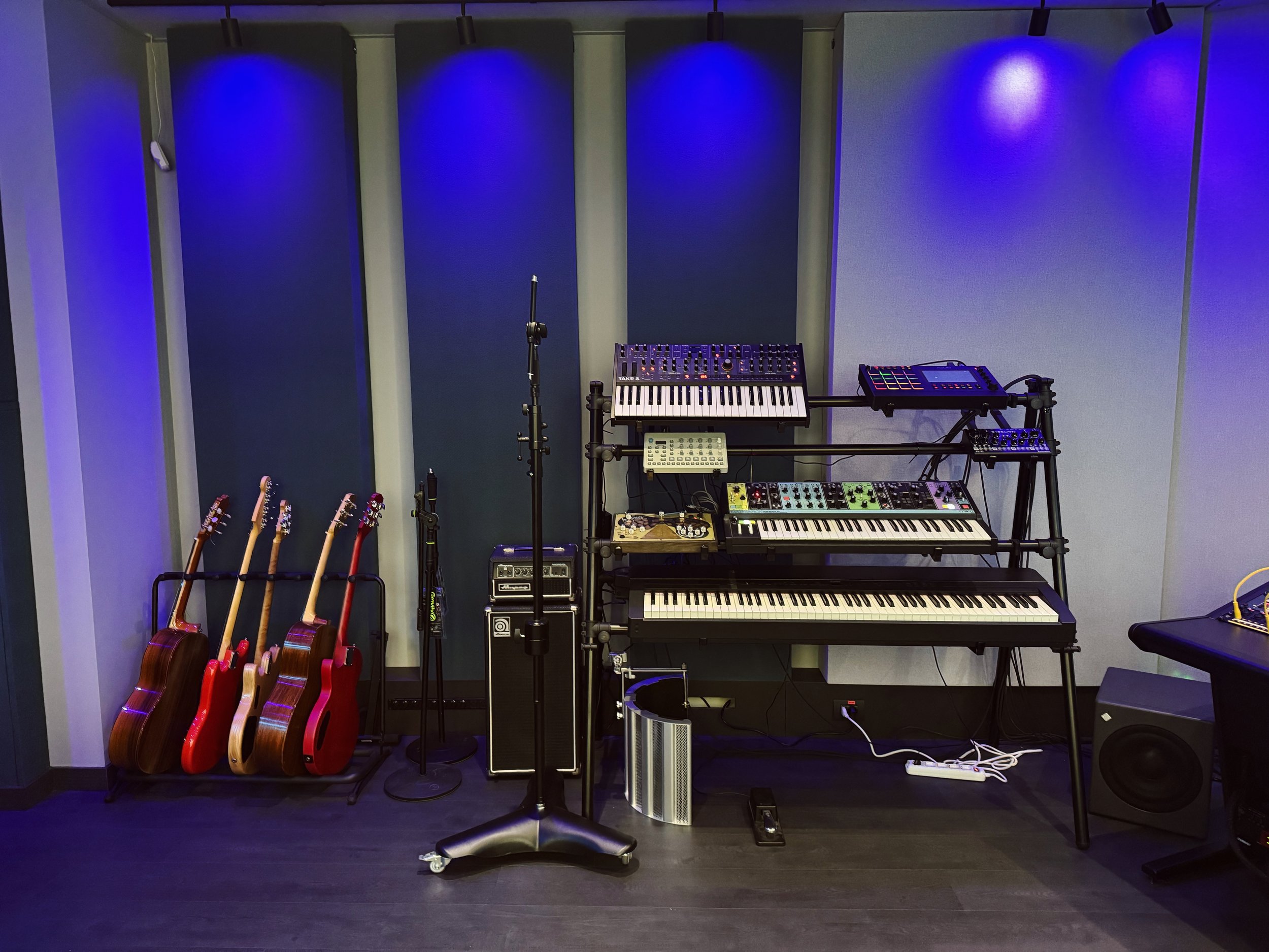 Stanza con strumenti musicali, incluso chitarre appoggiate a un supporto, una tastiera, un mixer, un sintetizzatore, un campionatore, e vari effetti e amplificatori, con pannelli acustici blu sullo sfondo.