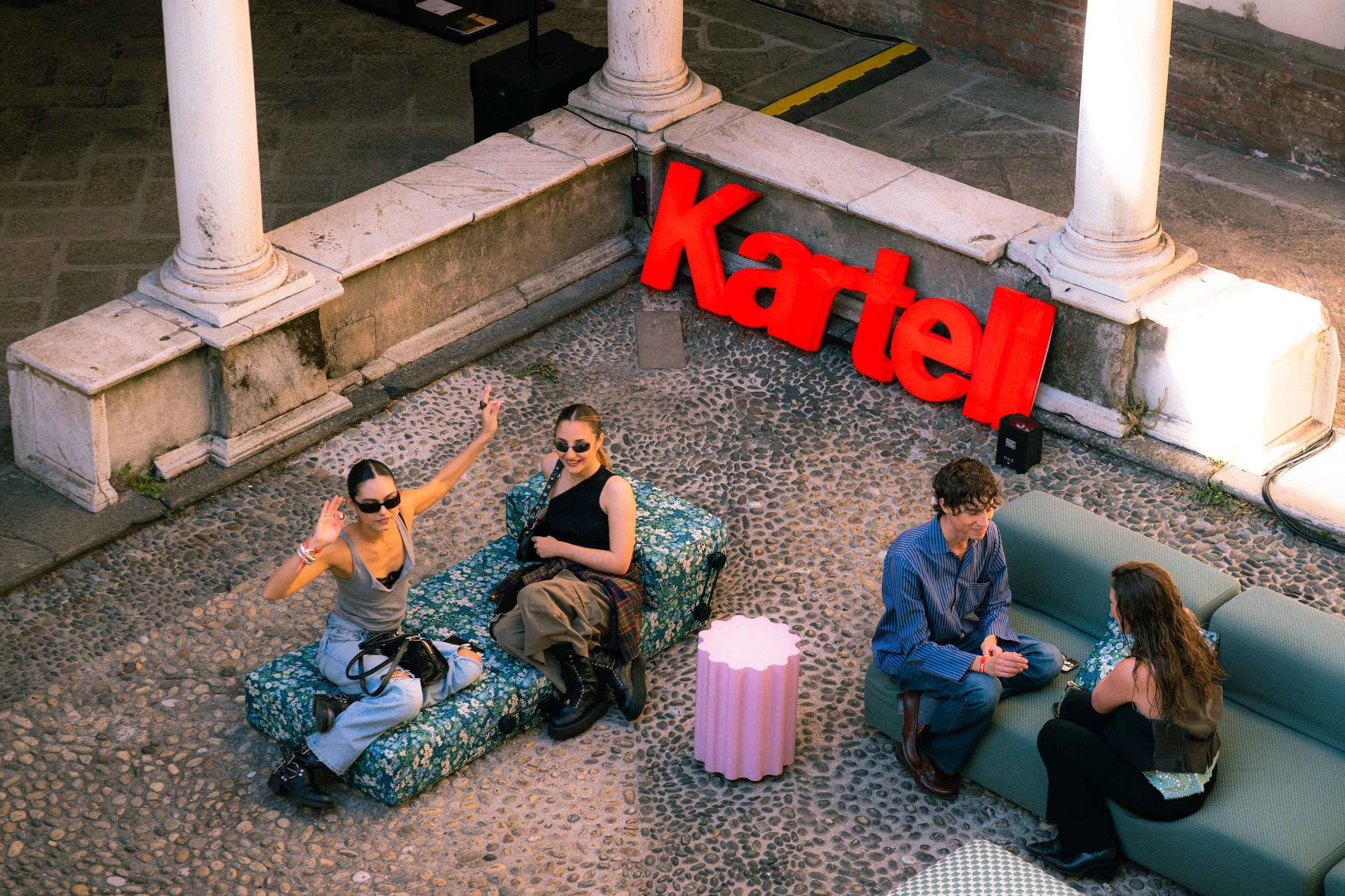 Gruppo di persone sedute e in piedi in un'area all'aperto, con alcuni che sorridono e fanno saluti. Sullo sfondo, un segno rosso gigante con la parola 'Karte' e colonne in marmo.