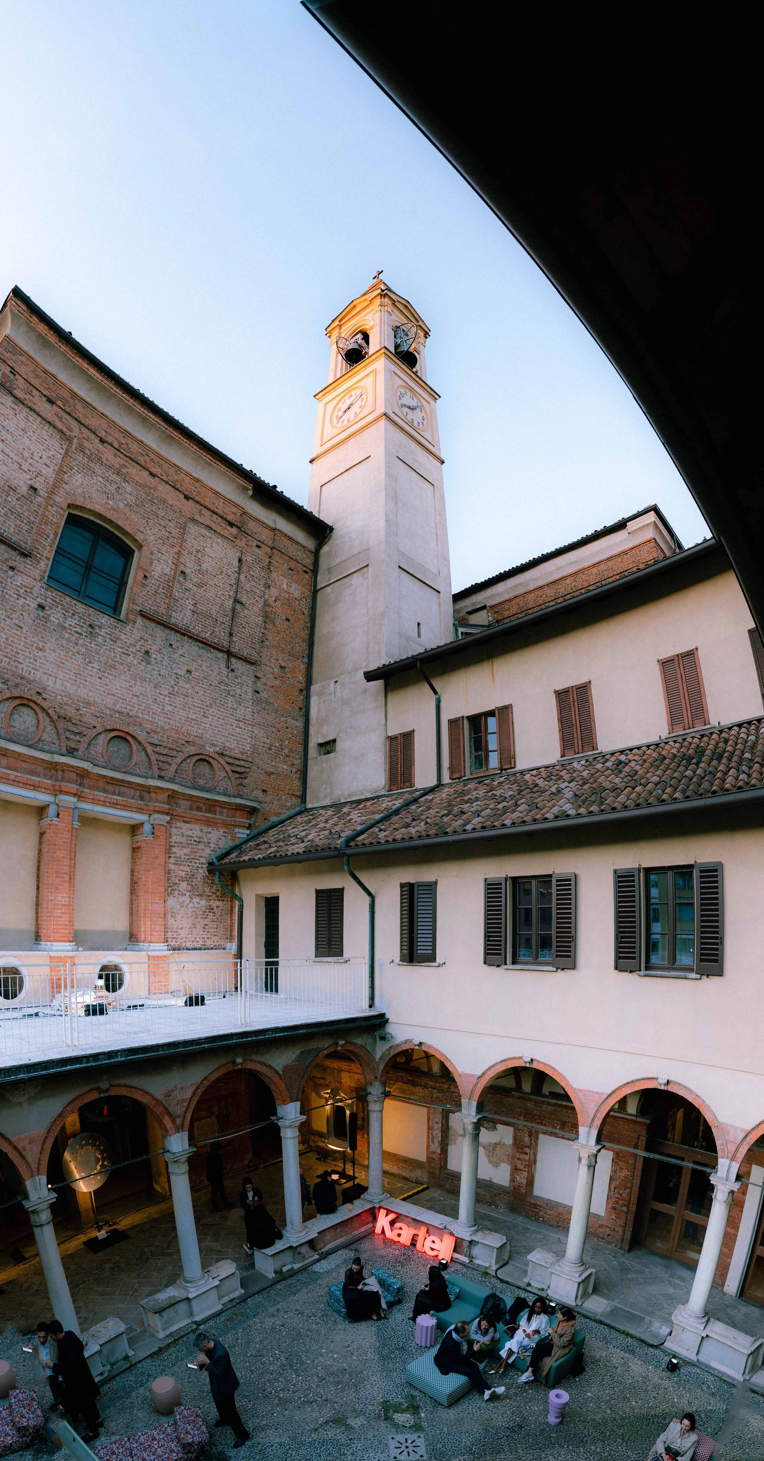 Cortile interno di un edificio storico con colonne e arcate, con persone sedute e in piedi, alcune che usano il telefono. Sullo sfondo, un'area con un'illuminazione rossa con il testo "Katel".