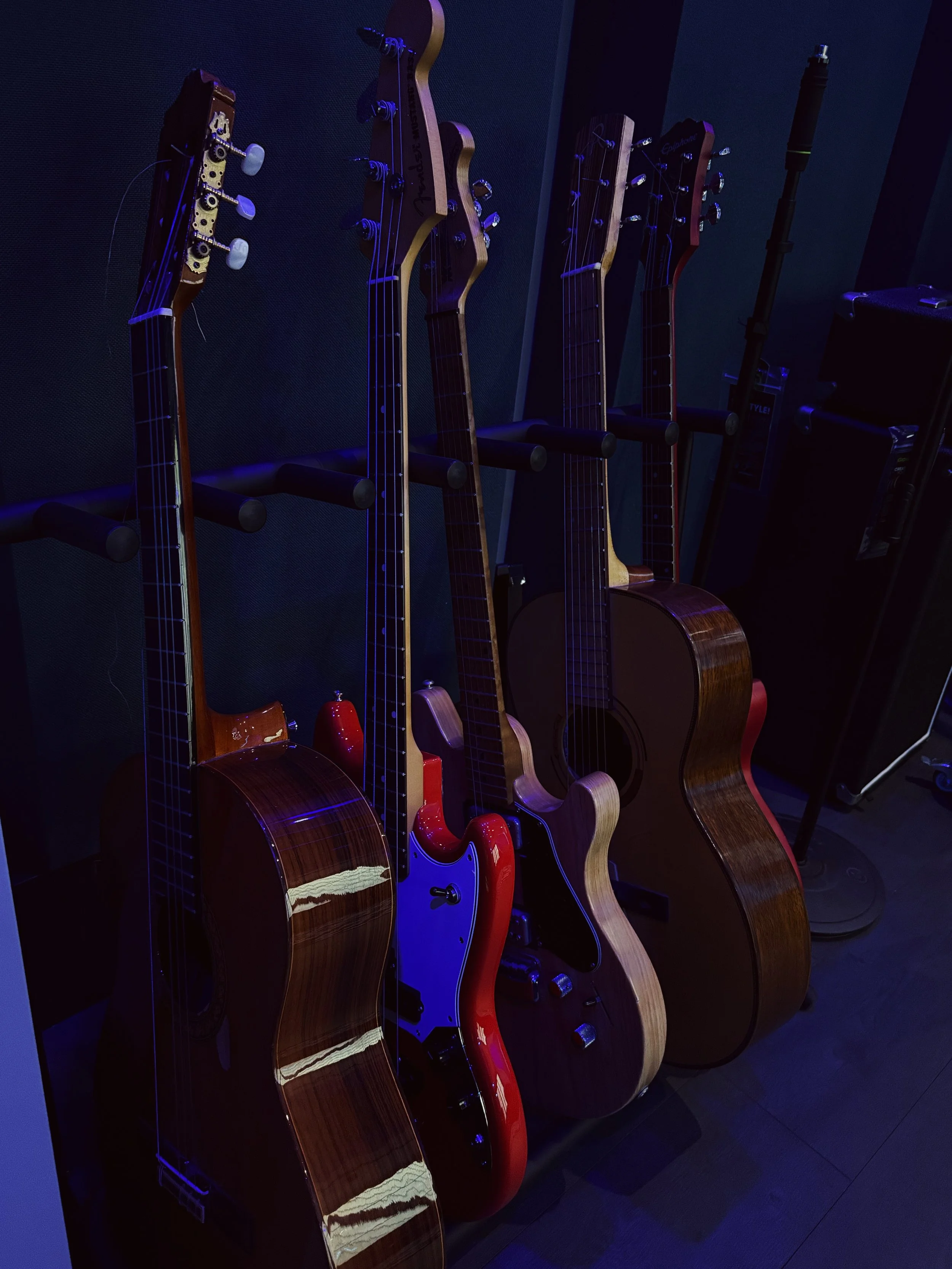 Cinque chitarre acustiche appoggiate a una parete con sfondo nero, illuminate da luci blu, in un ambiente probabilmente uno studio di registrazione o una sala prove.
