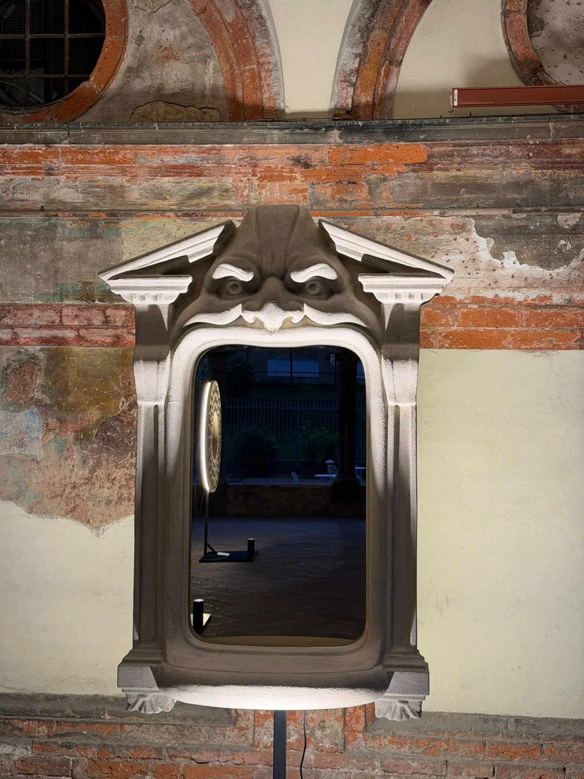 Specchio con cornice decorativa a forma di volto grottesco, posizionato di fronte a un muro di mattoni rossi e cemento.