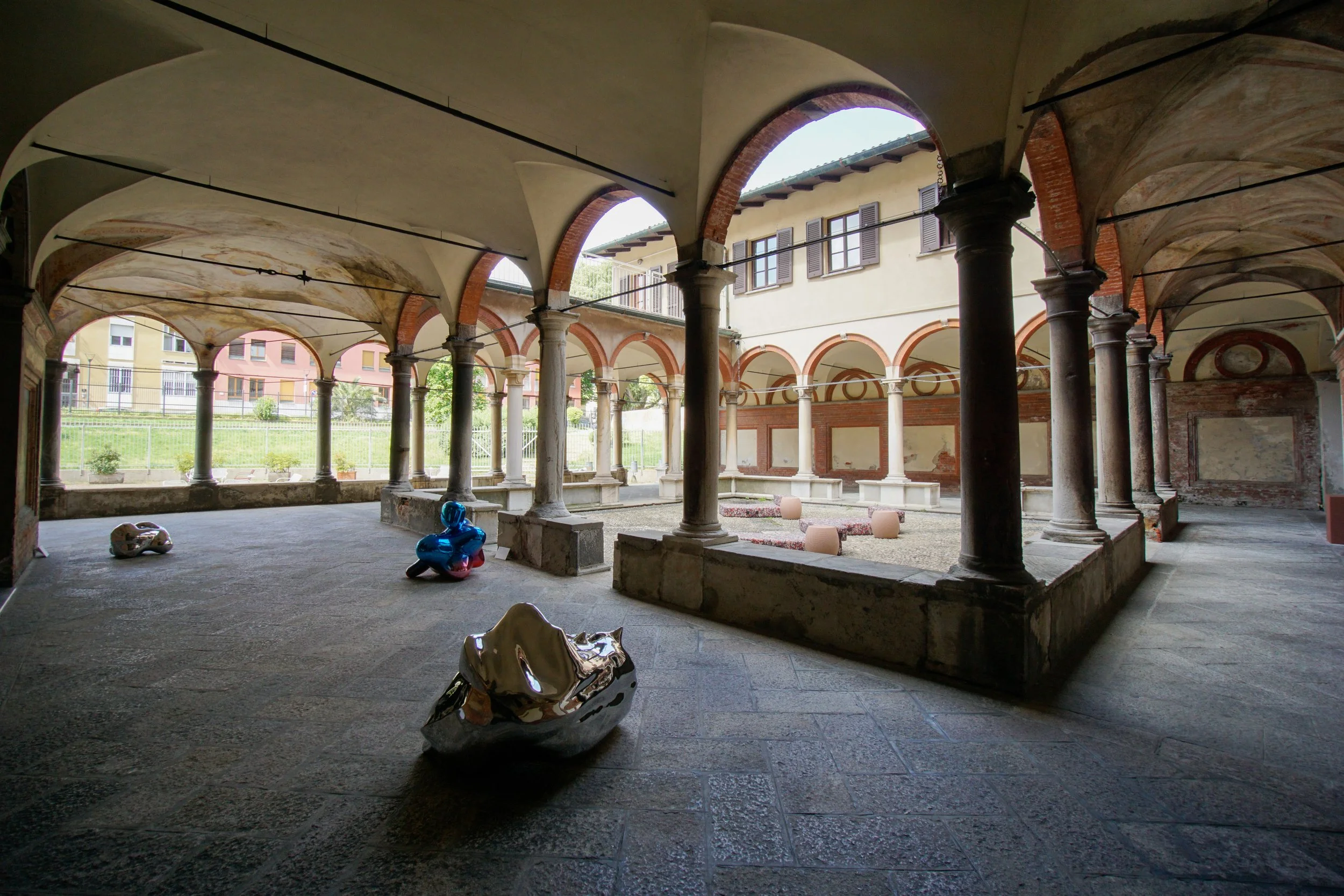 Cortile coperto con archi e colonne, con statue moderne in primo piano e edifici residenziali sullo sfondo.