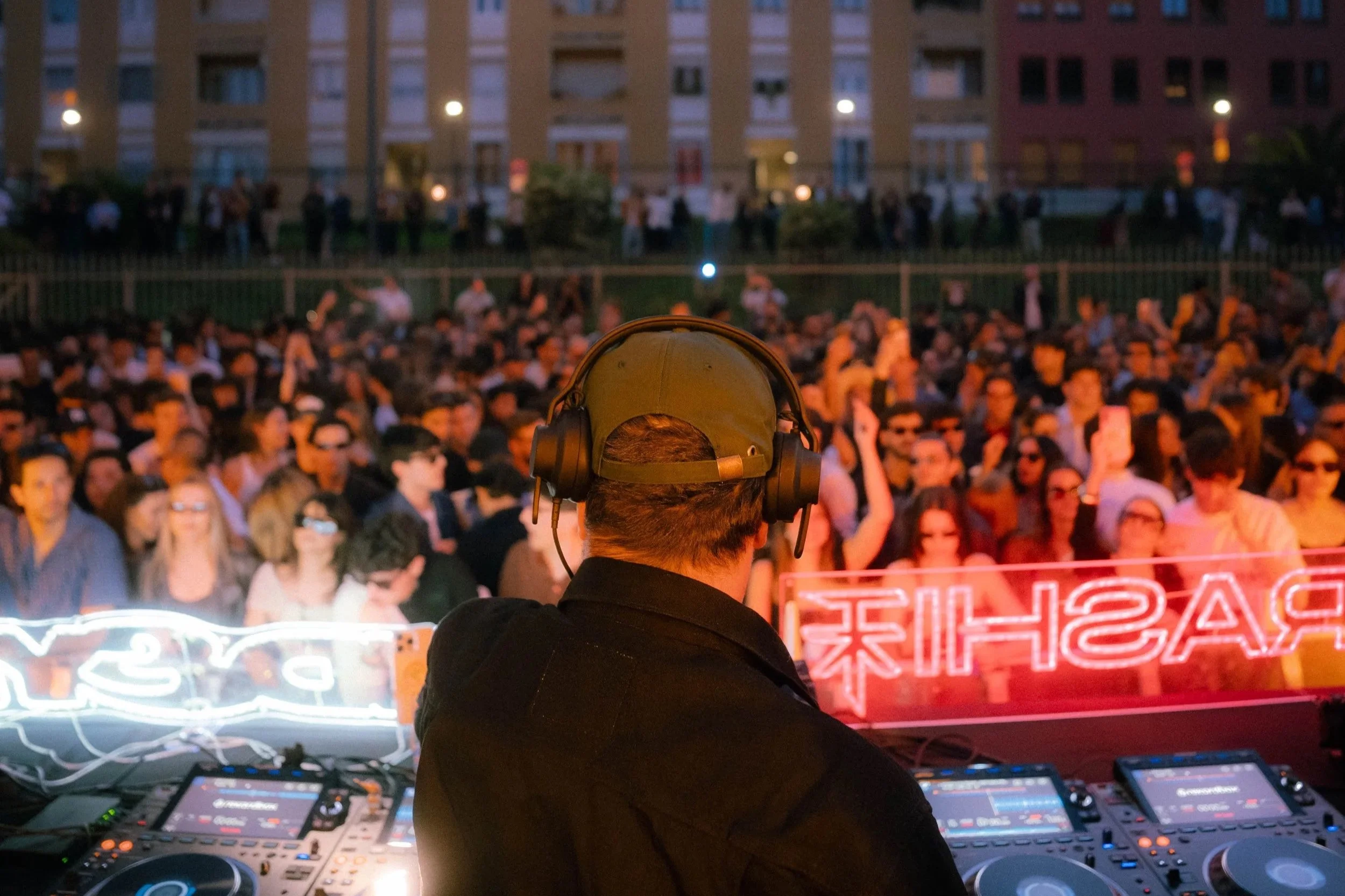 Un DJ con cuffie sopra la testa lavora con giradischi e controller DJ durante un evento all'aperto con molte persone che ascoltano e si divertono, in uno scenario cittadino di sera.