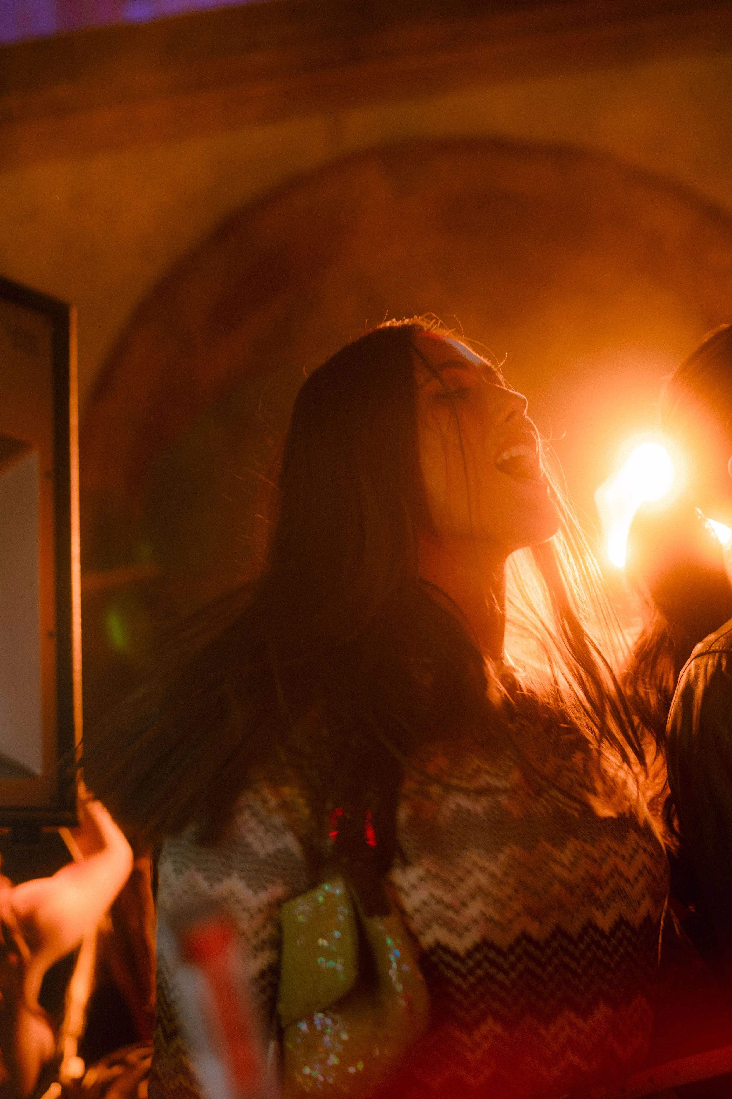 Una donna con capelli lunghi e scuri sorride e canta, illuminata da una luce calda proveniente da destra, in un ambiente buio.