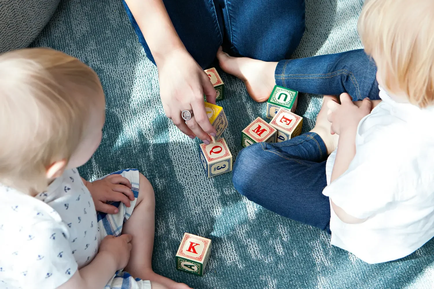 Two young children and an adult sitting on a blue carpet playing with colorful alphabet blocks.