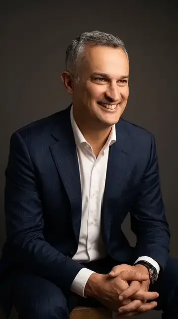 A smiling man in a navy suit and white shirt, sitting with his hands clasped, against a dark background.
