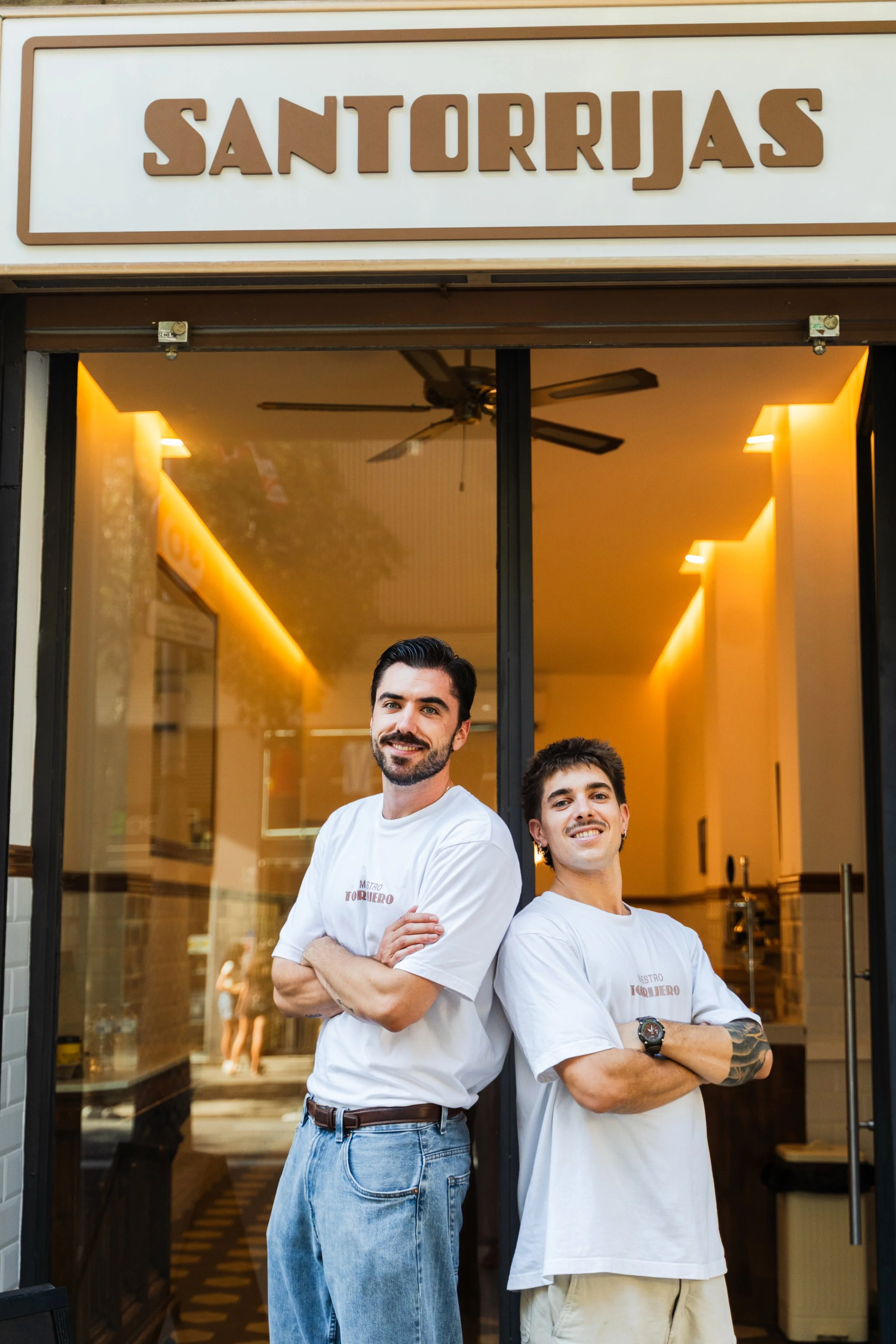 Dos jóvenes hombres con camisetas blancas con el logo de un restaurante llamado 'Nuestro Torillero', uno de ellos con cabello oscuro y barba, el otro más joven con tatuajes en brazos. Están de pie en la entrada de un establecimiento con una sonrisa confiada, cerca de una puerta de cristal con interior iluminado y decoración moderna.
