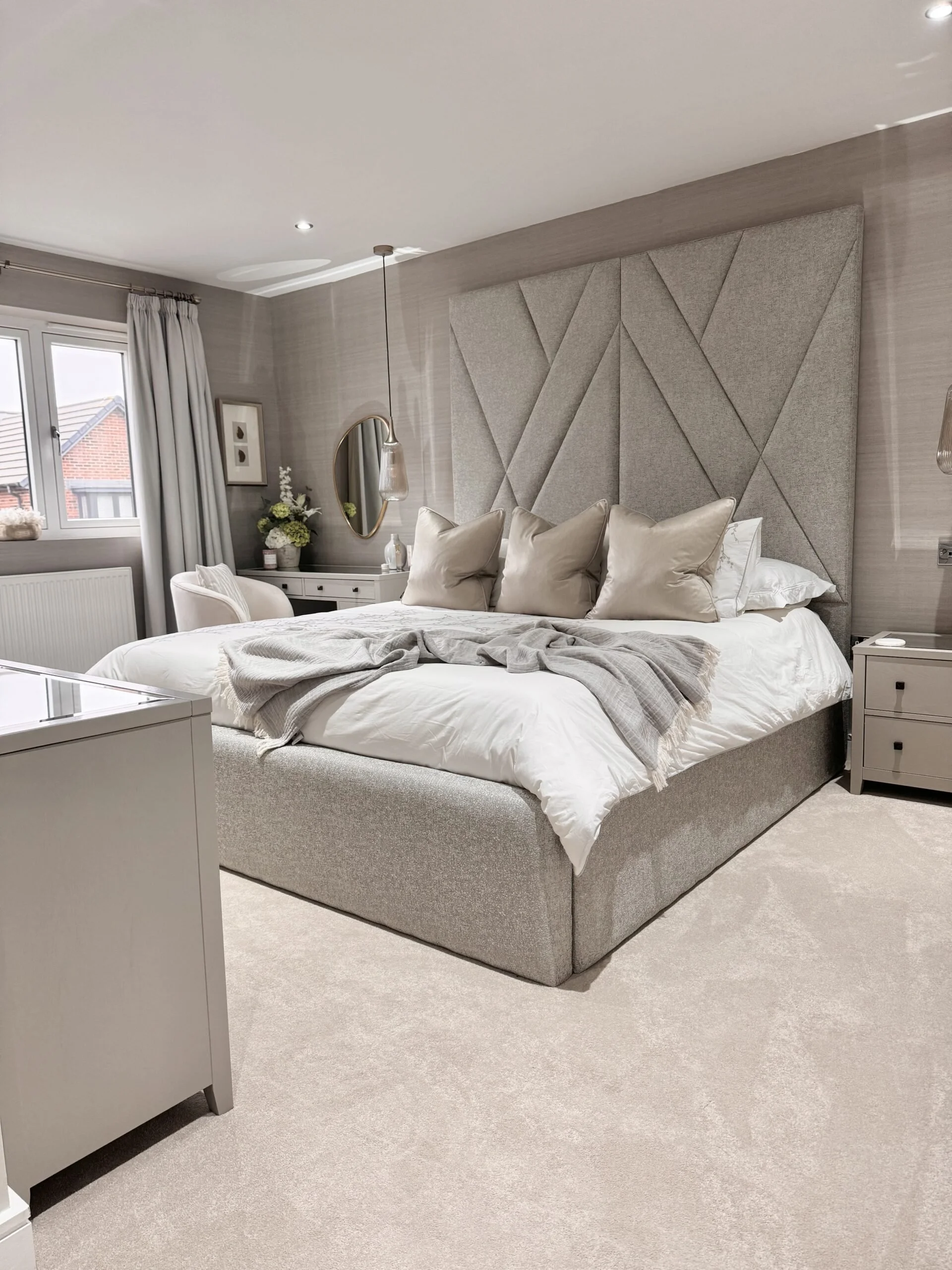 A modern bedroom with a large bed featuring a gray upholstered headboard, beige pillows, a white bedspread, and a gray throw blanket. The room has light gray walls, a window with white curtains, a white nightstand with a vase of flowers, a mirror, and a pendant light hanging from the ceiling.