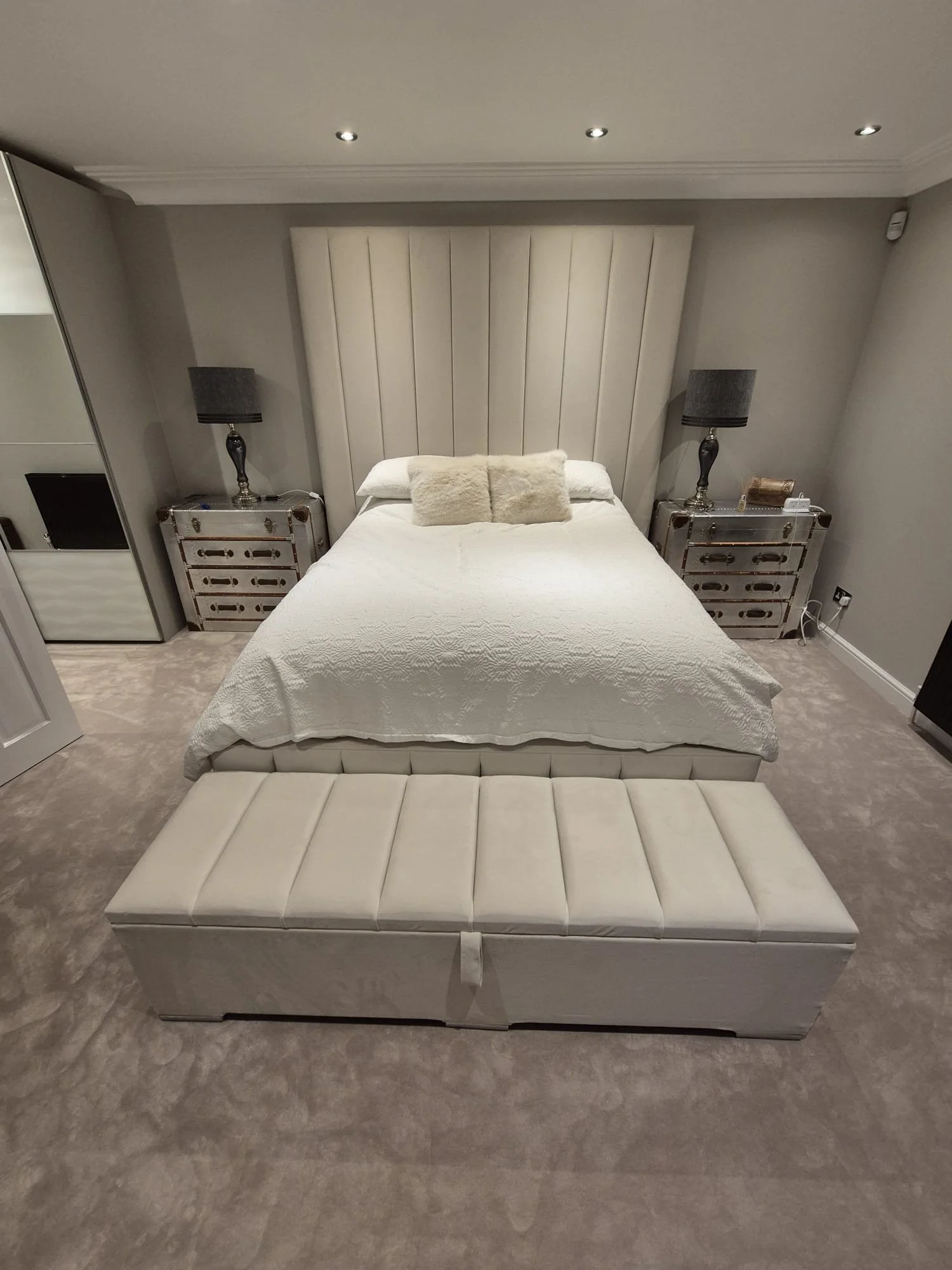 A neatly made bed with white bedding and two cream-colored pillows in a bedroom. Flanked by black lamps on decorative silver nightstands, with a tall upholstered cream headboard and gray walls.