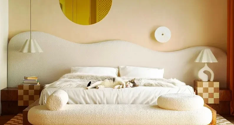 Modern bedroom with a cream-colored bed, a cat lying on the bed, nightstands with lamps, a round mirror, and minimalist decor.