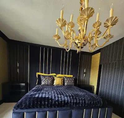 A bedroom with black walls, a dark bed with black and gold pillows, and a gold chandelier hanging from the ceiling.