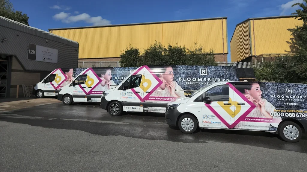 Four van vehicles parked in a lot with dental advertising wrap, featuring a woman with dark hair, a pink and gold diamond logo, and Bloomsbury Carpets & Flooring signage, near a large industrial building with a yellow end and trees.