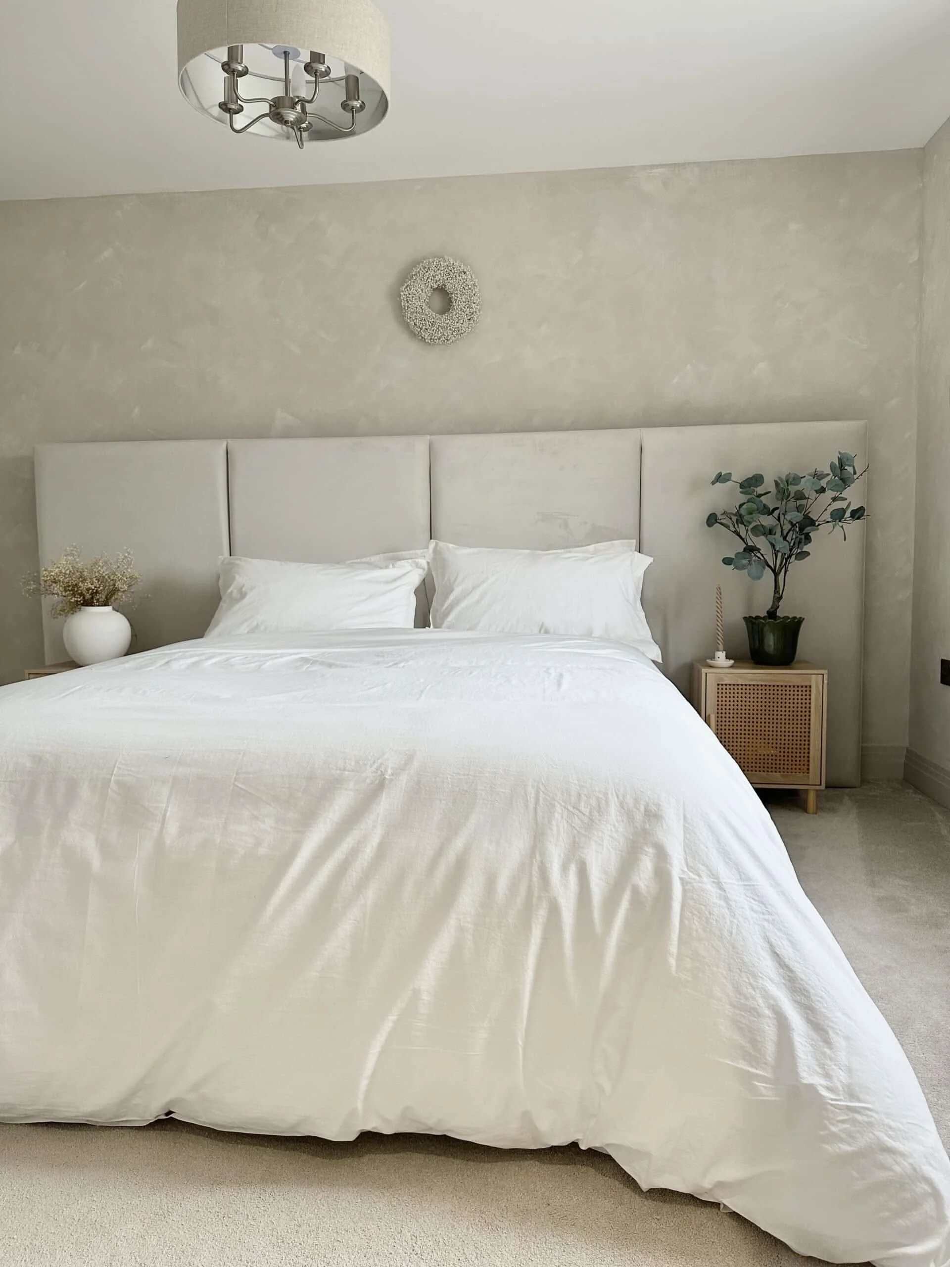 A bedroom with a white bed, white bedding, a beige headboard, a small wooden nightstand, a potted plant, and decorative wall art, with a ceiling light fixture.