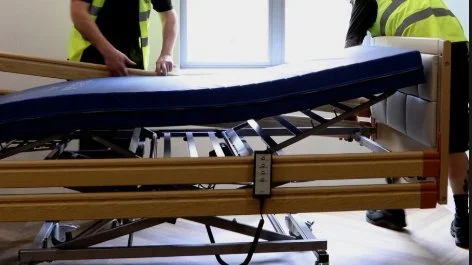 Two people in safety vests adjusting an empty hospital bed.