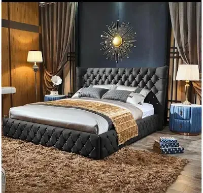 A modern bedroom with a tufted upholstered bed, gray bedding, and a decorative wall art piece featuring a sunburst design. There are two bedside tables with lamps, one blue with a side drawer and a white flower, and the other black. A brown area rug covers the wooden floor.