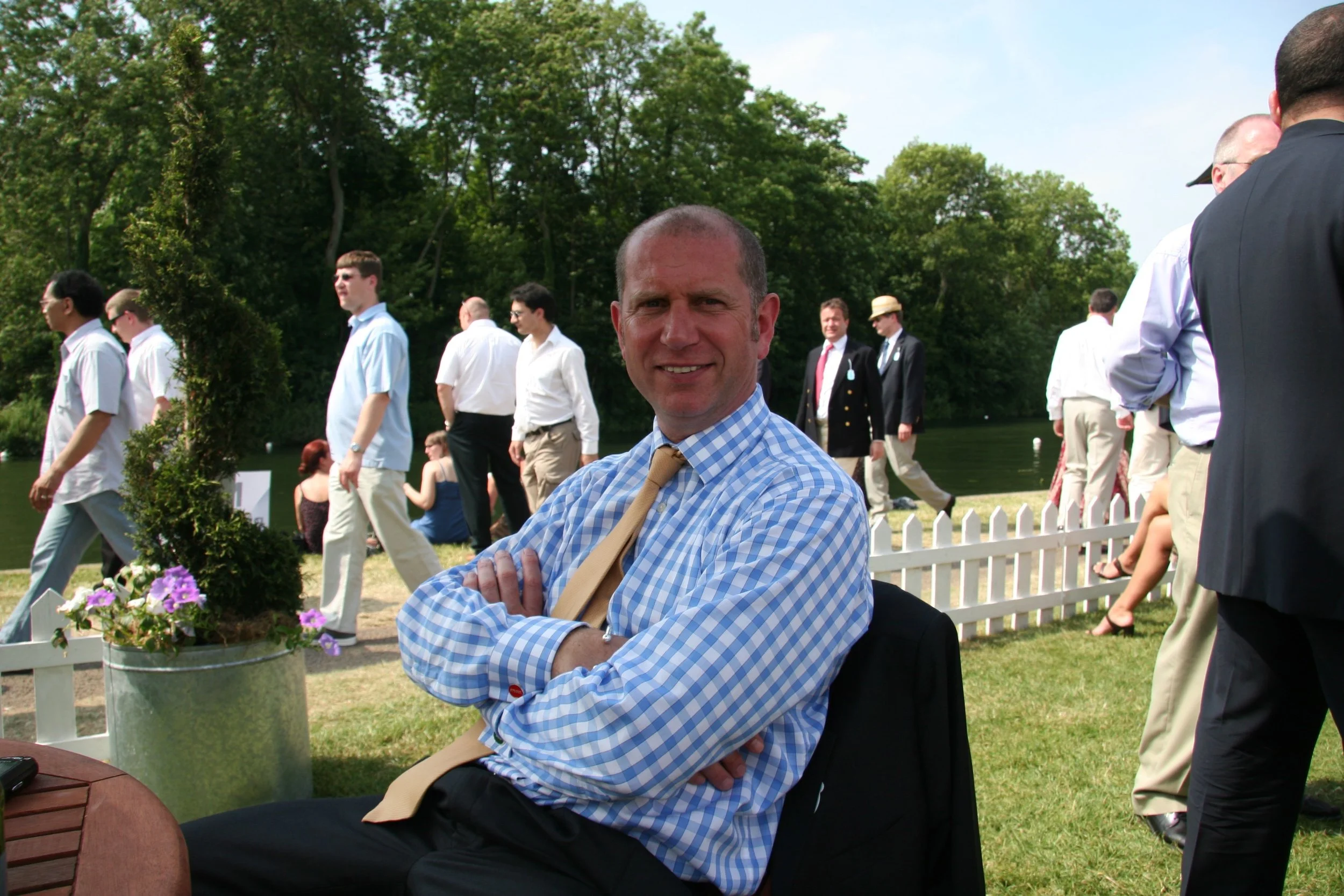 Man with crossed arms, smiling, wearing a blue checkered shirt and beige tie, sitting outdoors at a sunny event with other people walking and standing behind a white picket fence, trees in the background.