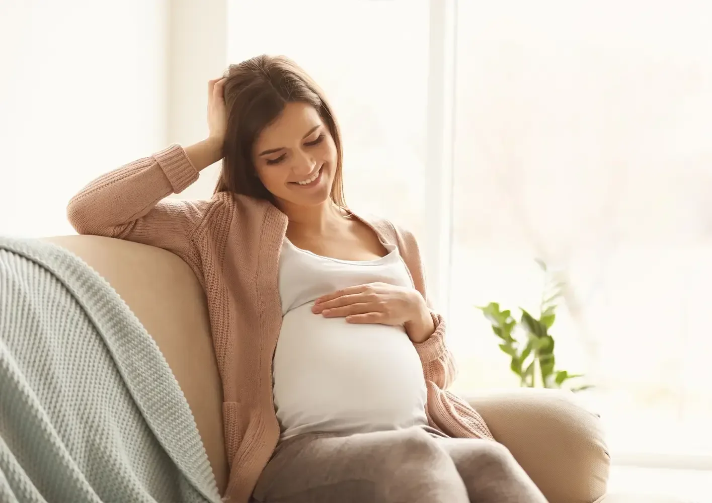 Pregnant woman sitting on a beige couch, smiling and touching her belly with one hand, in a bright room with natural light and a plant in the background.
