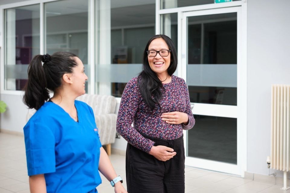 A woman in a blue medical scrub talking to a woman with glasses, long black hair, and a floral blouse, both smiling and engaging in conversation indoors.