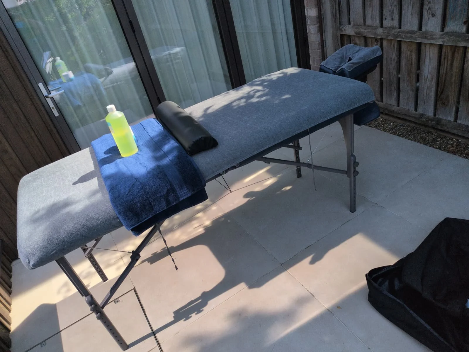 Outdoor massage table with a gray cover and blue towel, set on a patio with a wooden fence and sliding glass door. A yellow massage oil bottle sits on the towel.