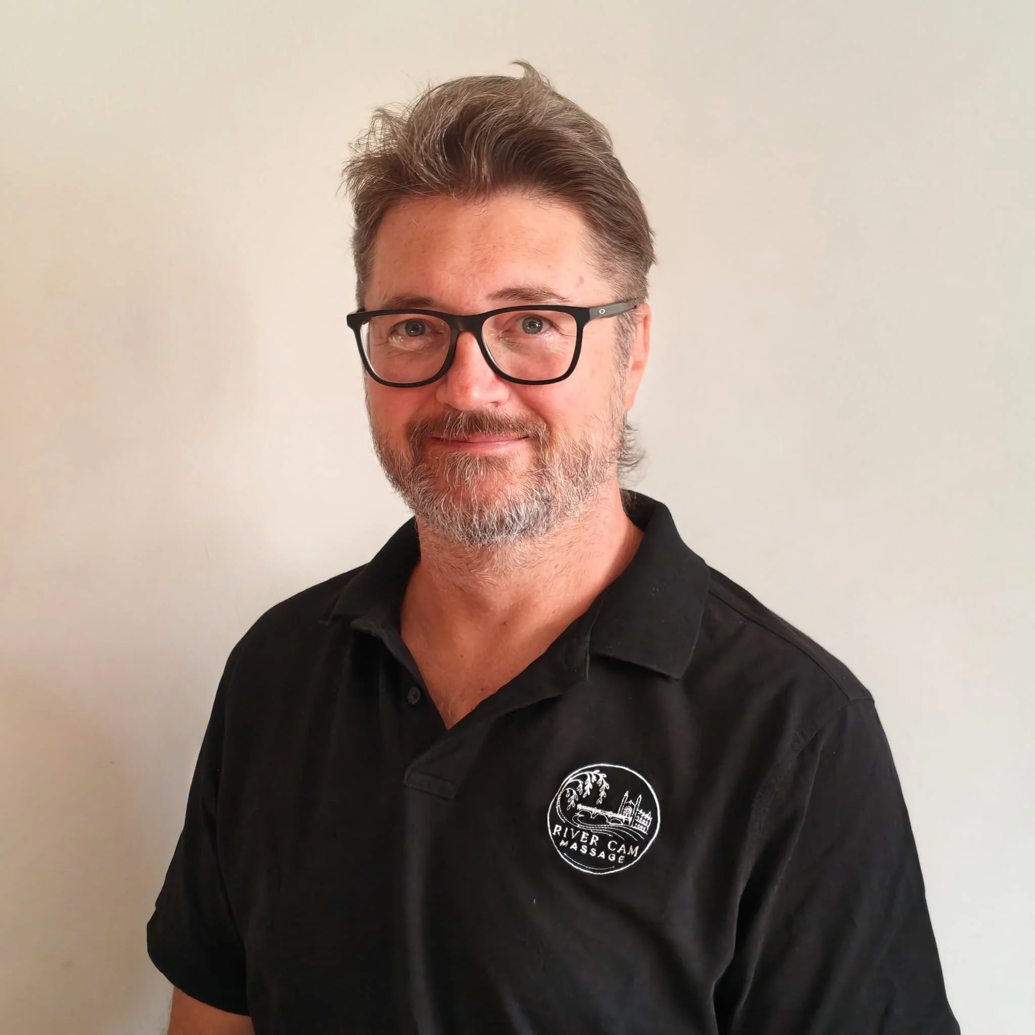 A man with dark brown hair and glasses, wearing a black shirt with a logo that reads "River Cam Massage," standing against a plain light-colored wall.