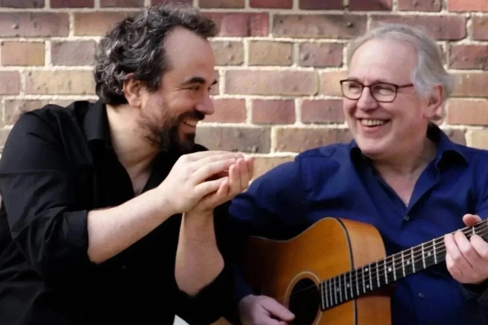 Zwei Männer lachen und spielen Gitarre vor einer Ziegelwand, einer hält die Hand des anderen.