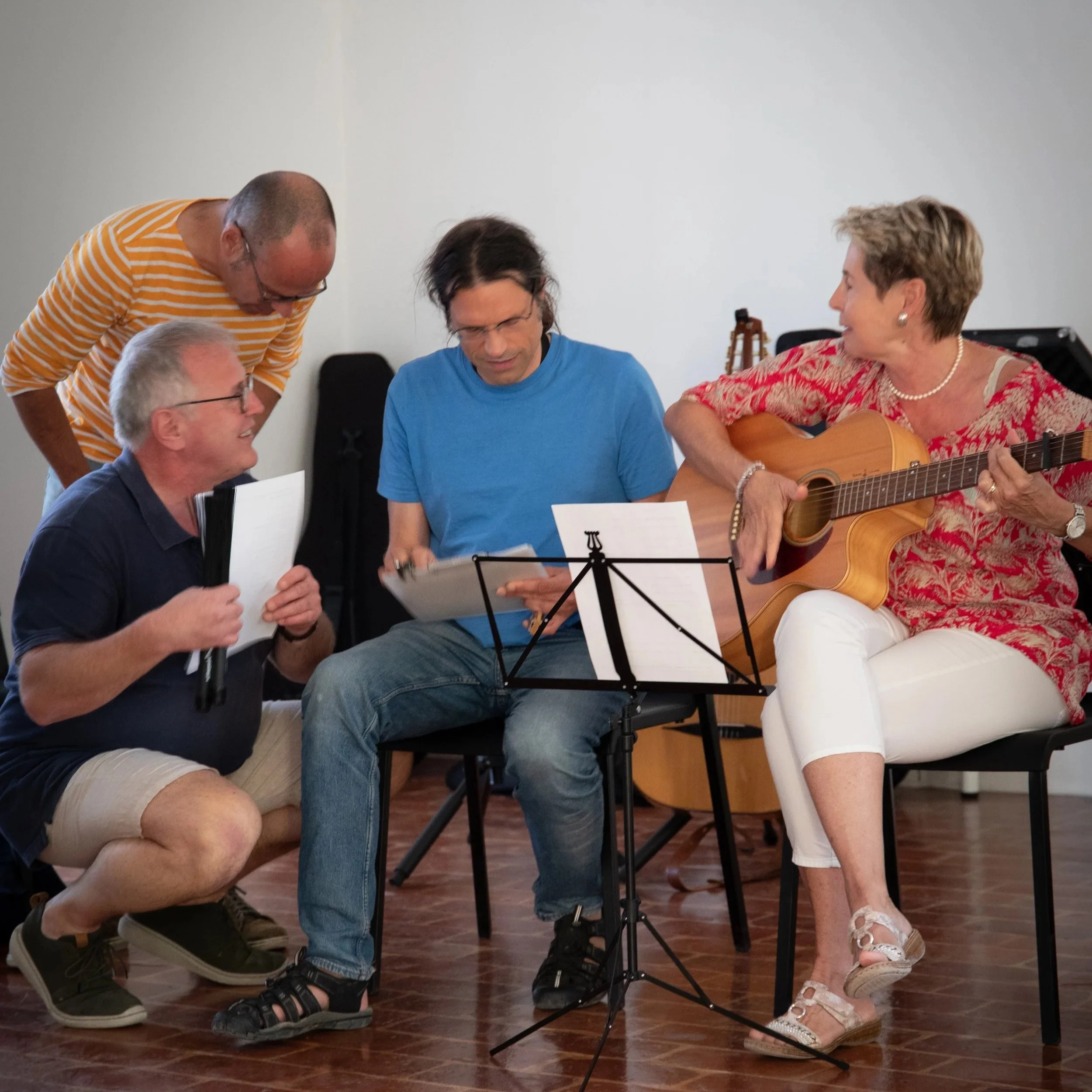 Fünf Menschen sitzen und stehen um ein Notenblatt, eine Frau spielt Gitarre, drei Männer schauen und singen, in einem Raum mit Holzboden.