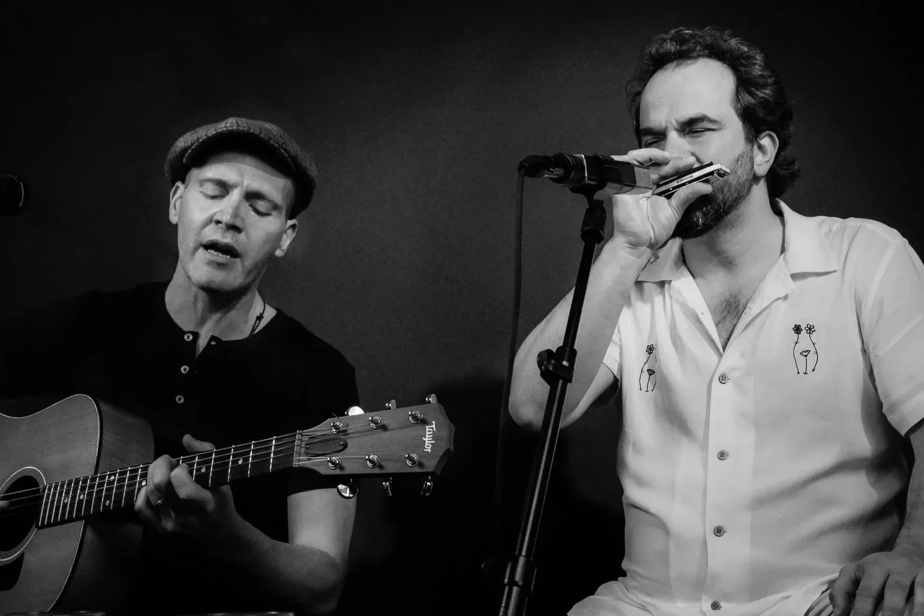 Zwei Männer, einer spielt Gitarre und der andere singt in einem Mikrofon, in Schwarz-Weiß aufgenommen.