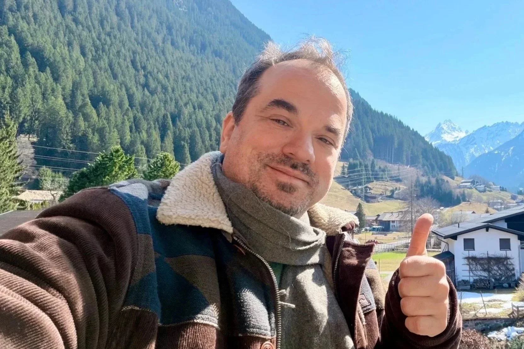 Ein Mann macht ein Selfie vor einer Berglandschaft mit Häusern, Bäumen und schneebedeckten Bergen im Hintergrund, zeigt Daumen nach oben.