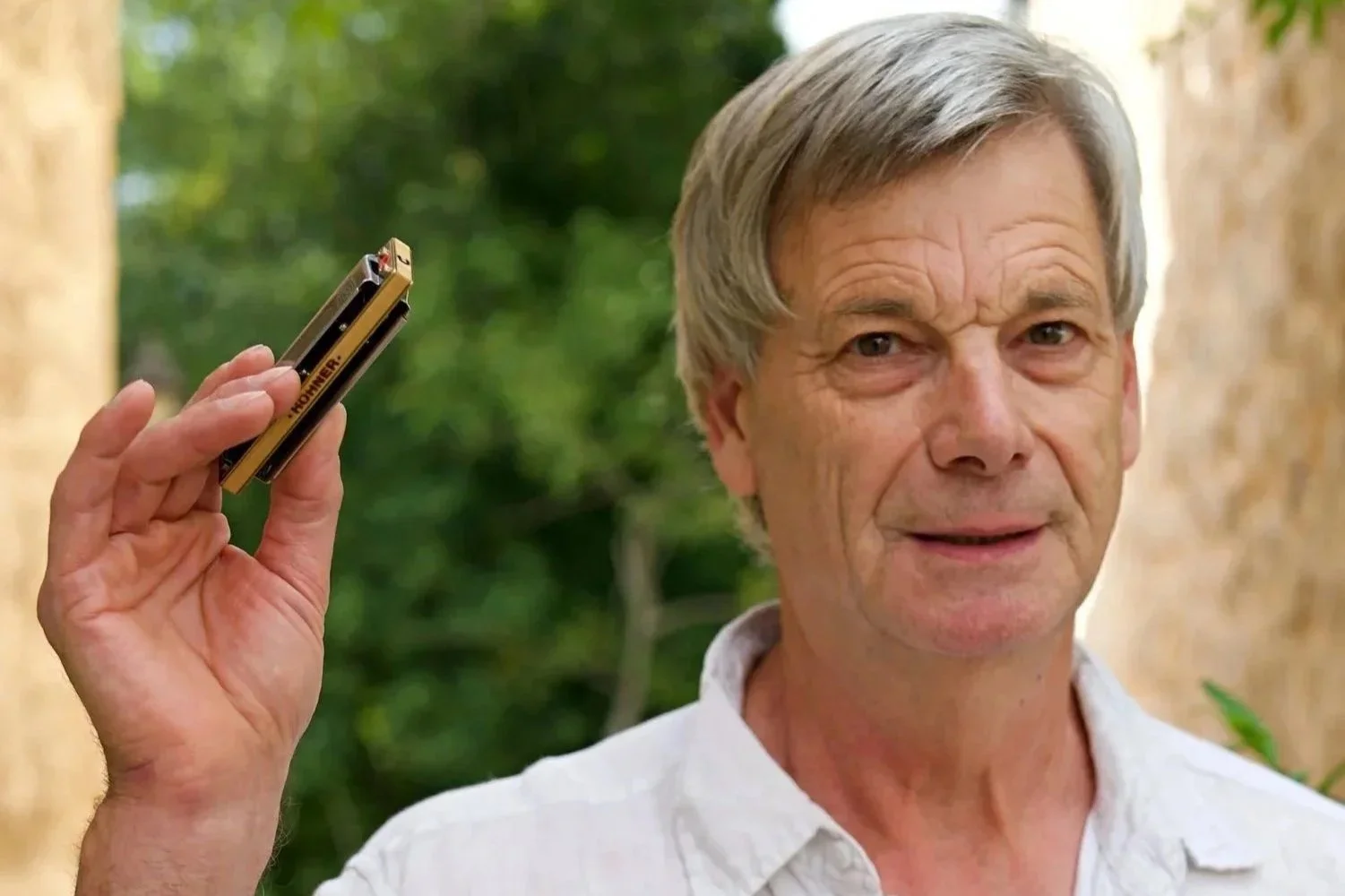 Ein älterer Mann mit grauem Haar hält ein kleines goldenes Werkzeug in der Hand, während er nach vorne schaut, im Hintergrund sind Bäume und eine Mauer zu sehen.