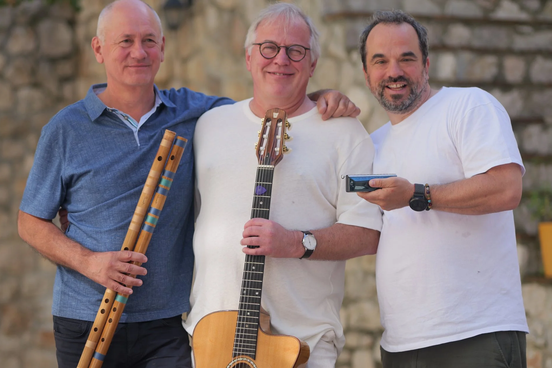 Drei Männer stehen eng beieinander, lächeln und zeigen ihre Musikinstrumente. Der Mann in der Mitte hält eine Gitarre, der Mann links hält zwei Flöten, und der Mann rechts hält ein Aufnahmegerät. Sie stehen vor einer Steinmauer im Freien.