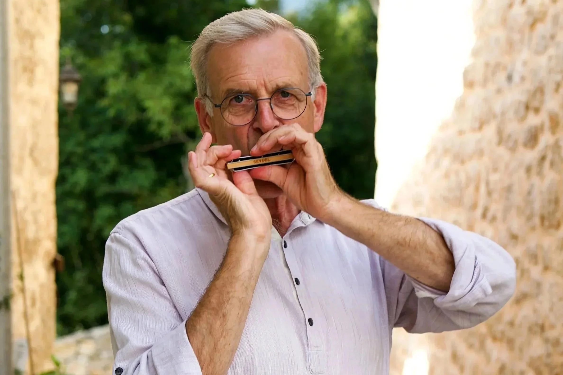 Älterer Mann mit Brille spielt Harmonica im Freien zwischen Bäumen.