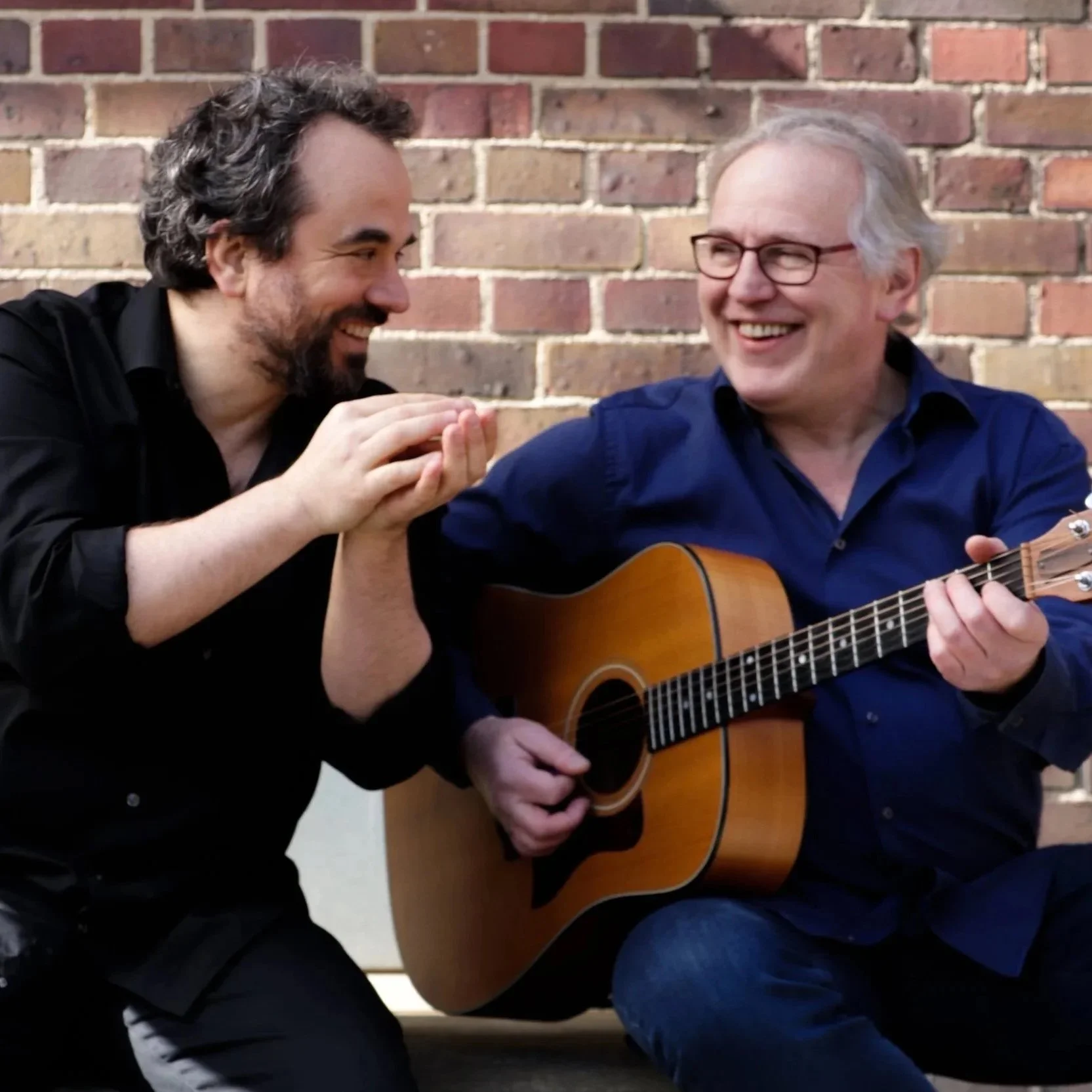 Lutz Schlosser und Marko Jovanović lachen beim Musizieren vor einer Ziegelmauer.